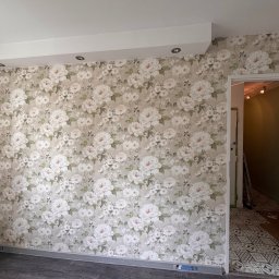 YEVHENII SEMENOV - Ściana z tapetą w kwiaty w jasnym pokoju. Widoczny fragment sufitu z oświetleniem punktowym i fragment podłogi. W tle widoczne wejście do innego pomieszczenia.