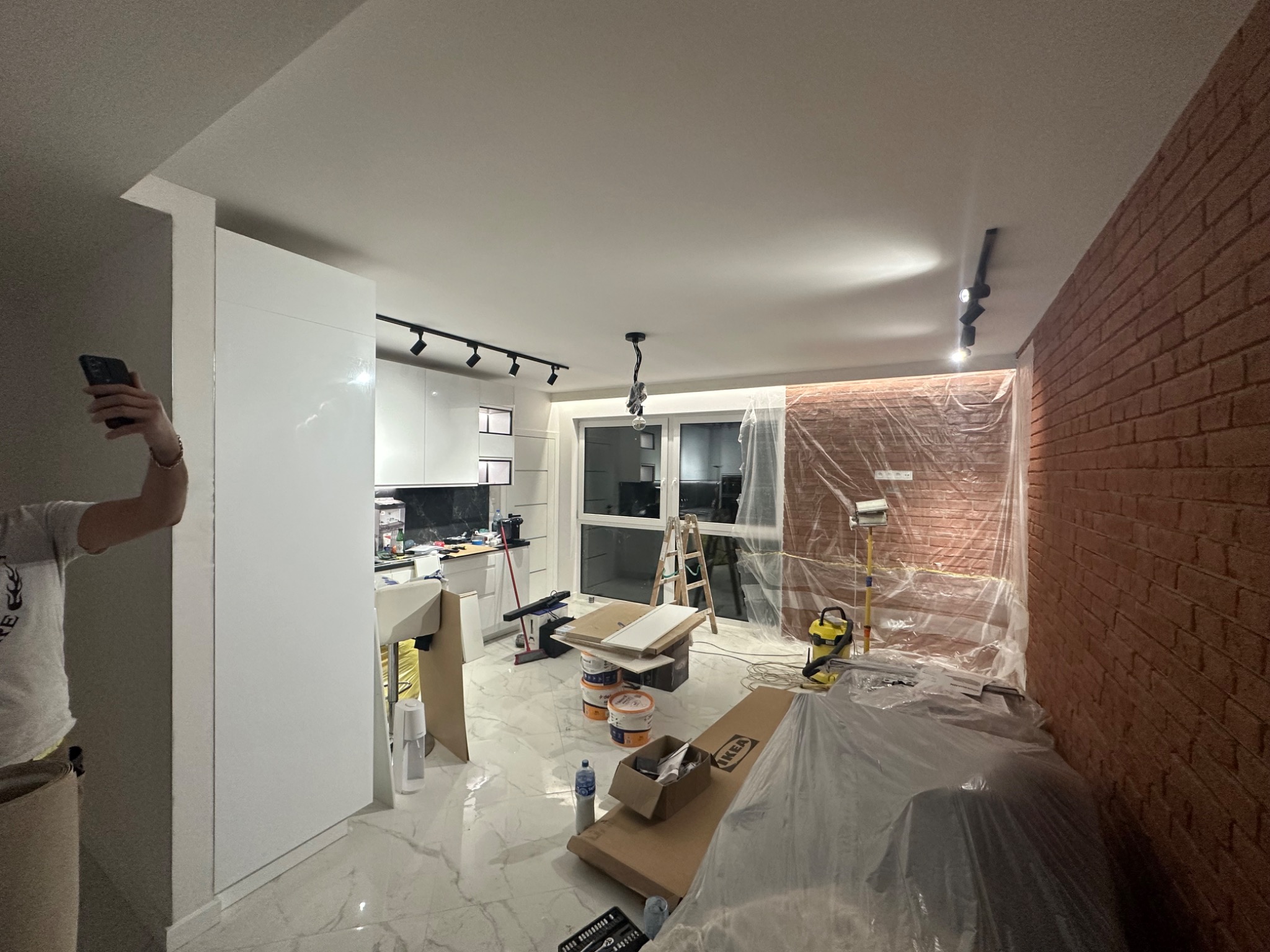 Wnętrze mieszkania w trakcie prac wykończeniowych: zabezpieczona folią ściana z cegły, widoczne puszki z farbą i kartony, białe meble kuchenne i marmurowe płytki podłogowe, osoba robiąca zdjęcie...