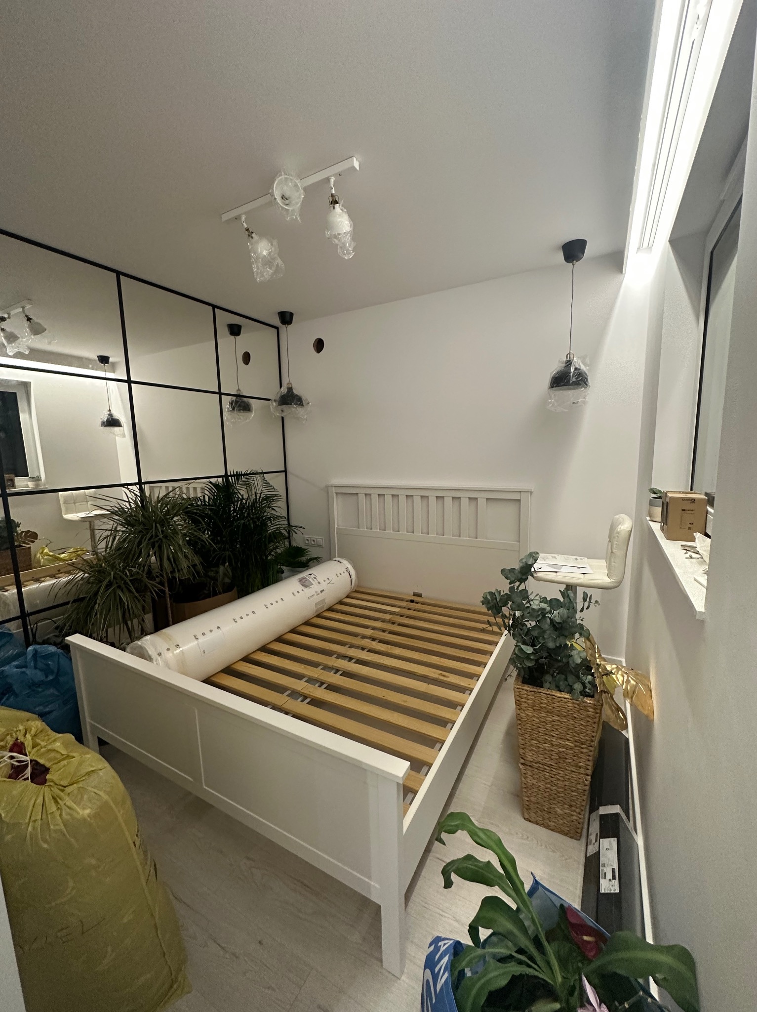 Białe łóżko z drewnianym stelażem, materac w folii, rośliny doniczkowe, lampy w folii ochronnej, lustrzana ściana w pokoju podczas urządzania.