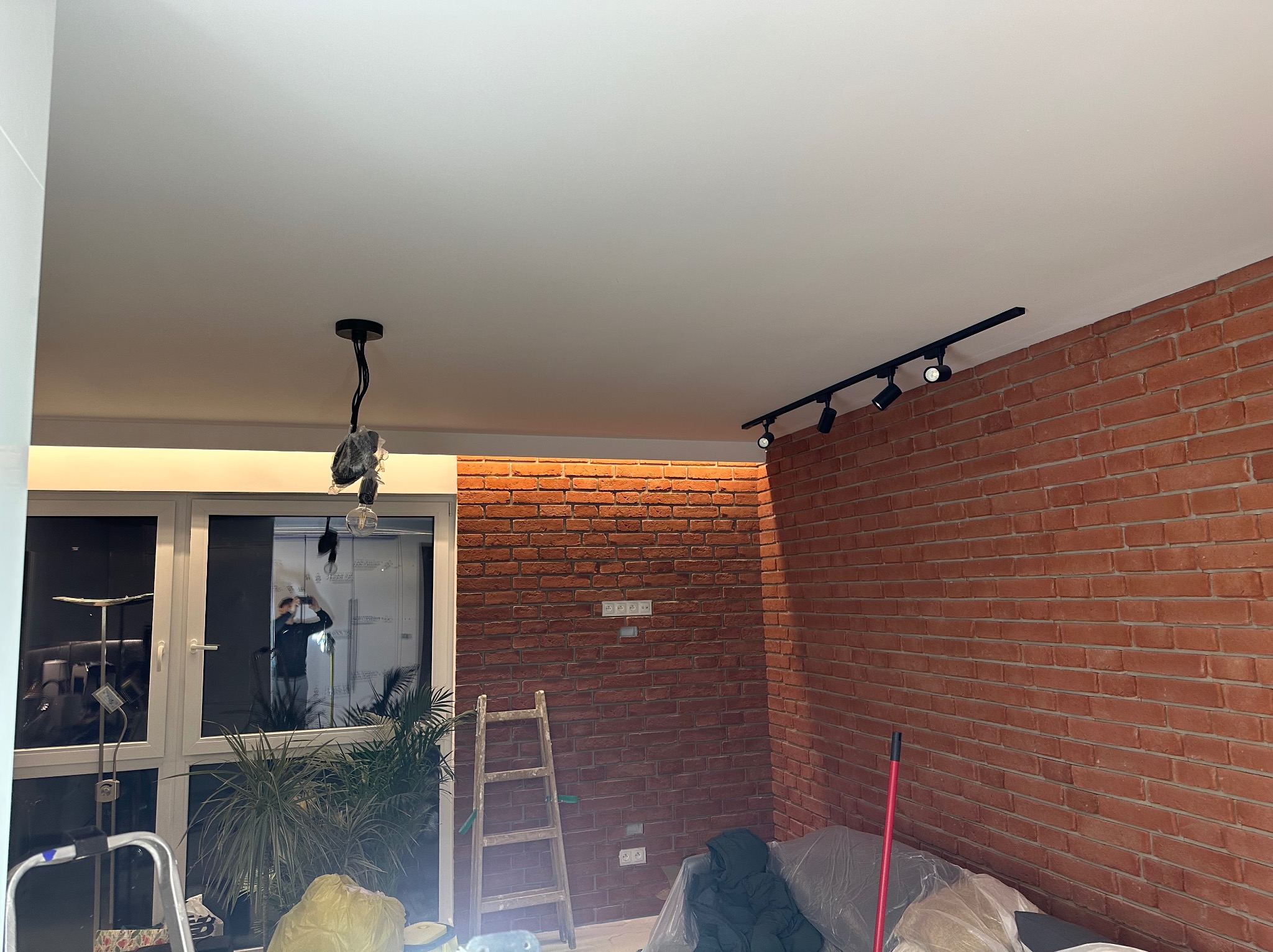 Wnętrze pokoju z czerwoną ceglaną ścianą, białym sufitem z czarnymi reflektorami, wiszącą lampą z niezabezpieczonymi kablami, drabiną opartą o ścianę i oknem z widoczną osobą robiącą zdjęcie.