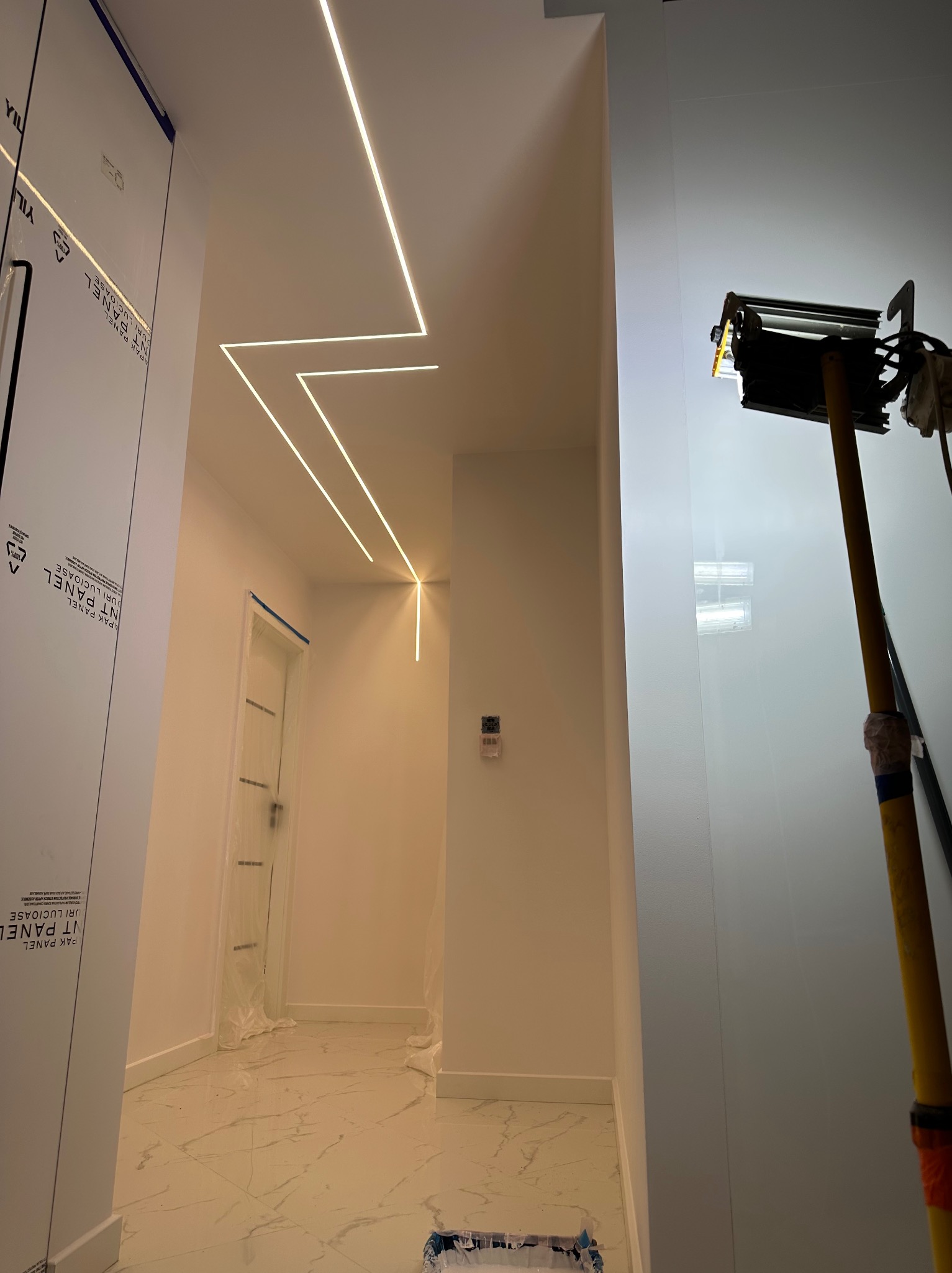 Korytarz w trakcie prac wykończeniowych z zamontowanym oświetleniem LED w suficie, zabezpieczona folią futryna drzwi, marmurowe płytki na podłodze i widoczna lampa budowlana na statywie.