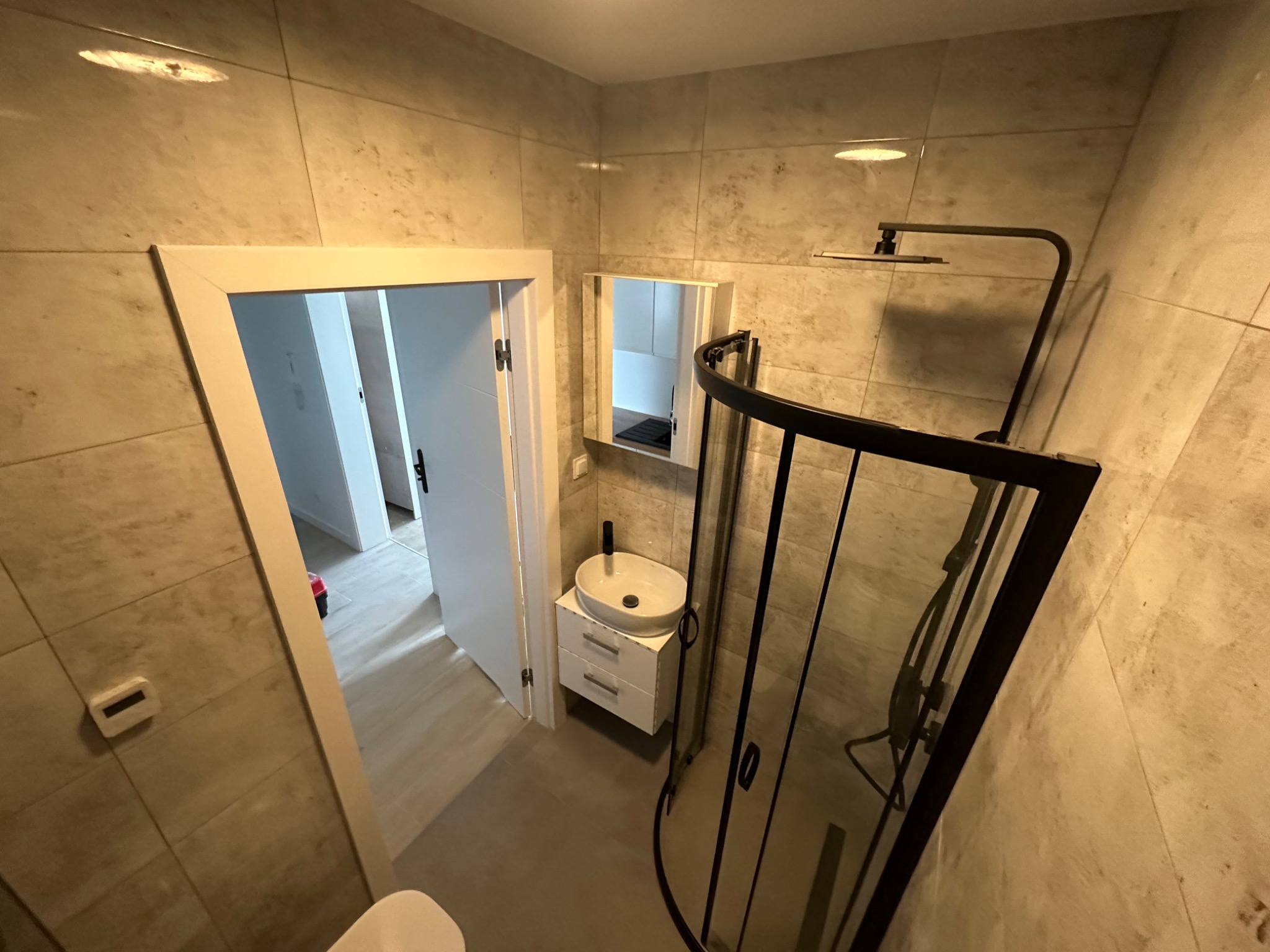 Nowoczesna łazienka z beżowymi płytkami, kabiną prysznicową z czarną ramą, umywalką nablatową na białej szafce i lustrem. Widoczny fragment toalety oraz otwarte drzwi do innego pomieszczenia.