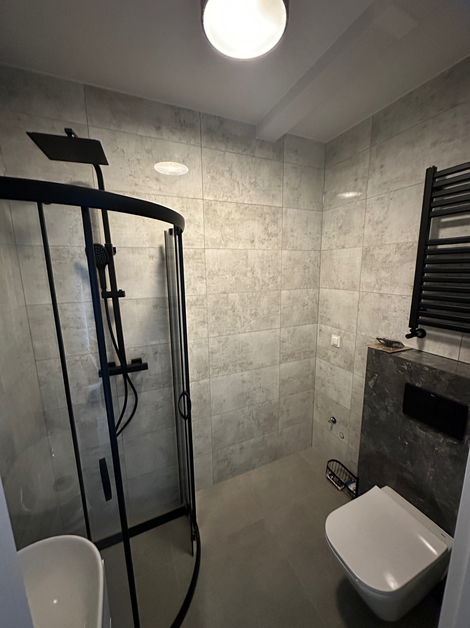 Nowoczesna łazienka z kabiną prysznicową typu walk-in z czarnymi ramami, wiszącą toaletą i grzejnikiem drabinkowym na tle szarych płytek imitujących kamień.