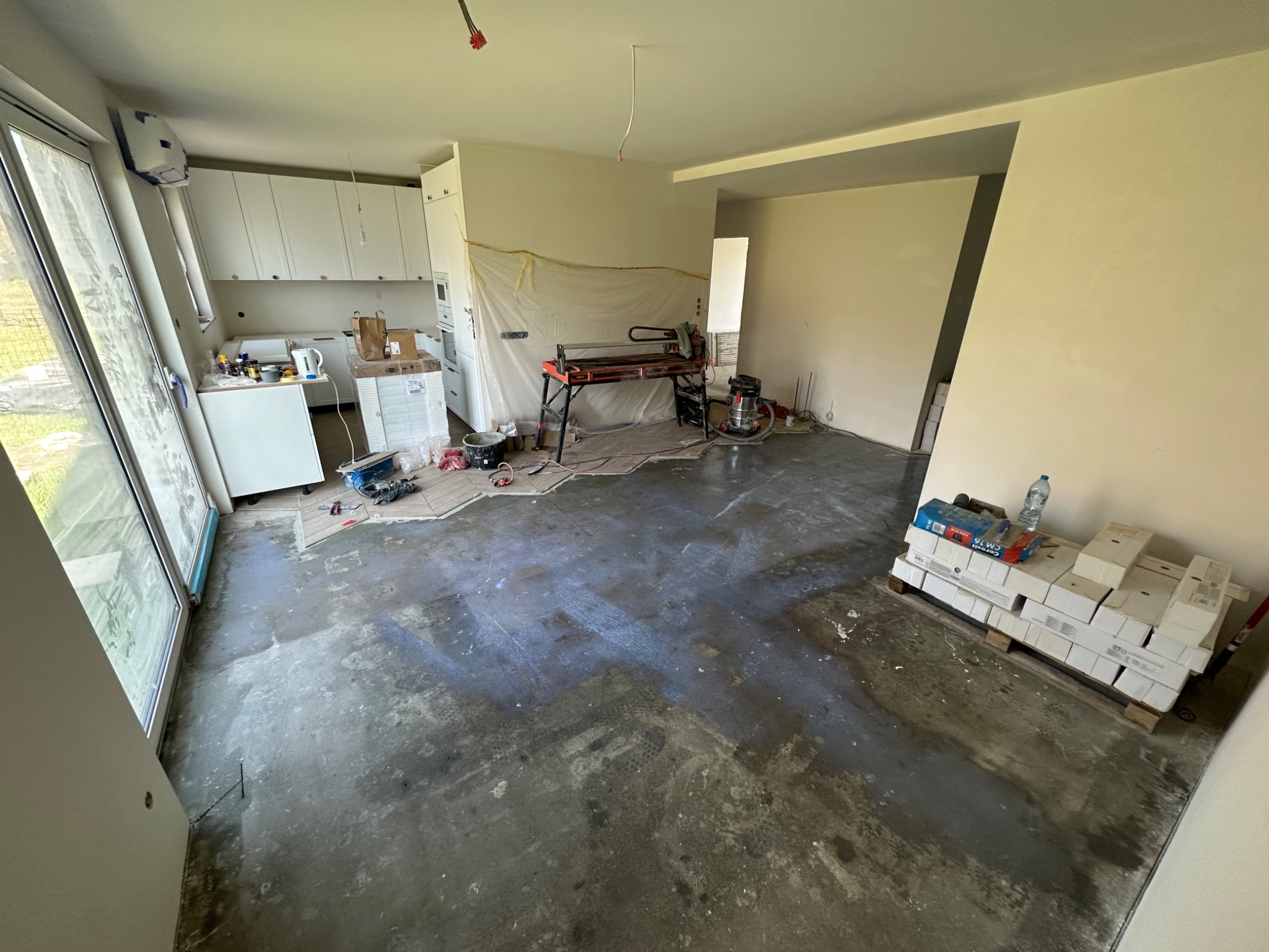 Wnętrze mieszkania w trakcie prac przygotowawczych do układania płytek, widoczna maszyna do cięcia płytek, paleta z opakowaniami płytek, częściowo ułożone płytki w kuchni, zakryte folią elementy.