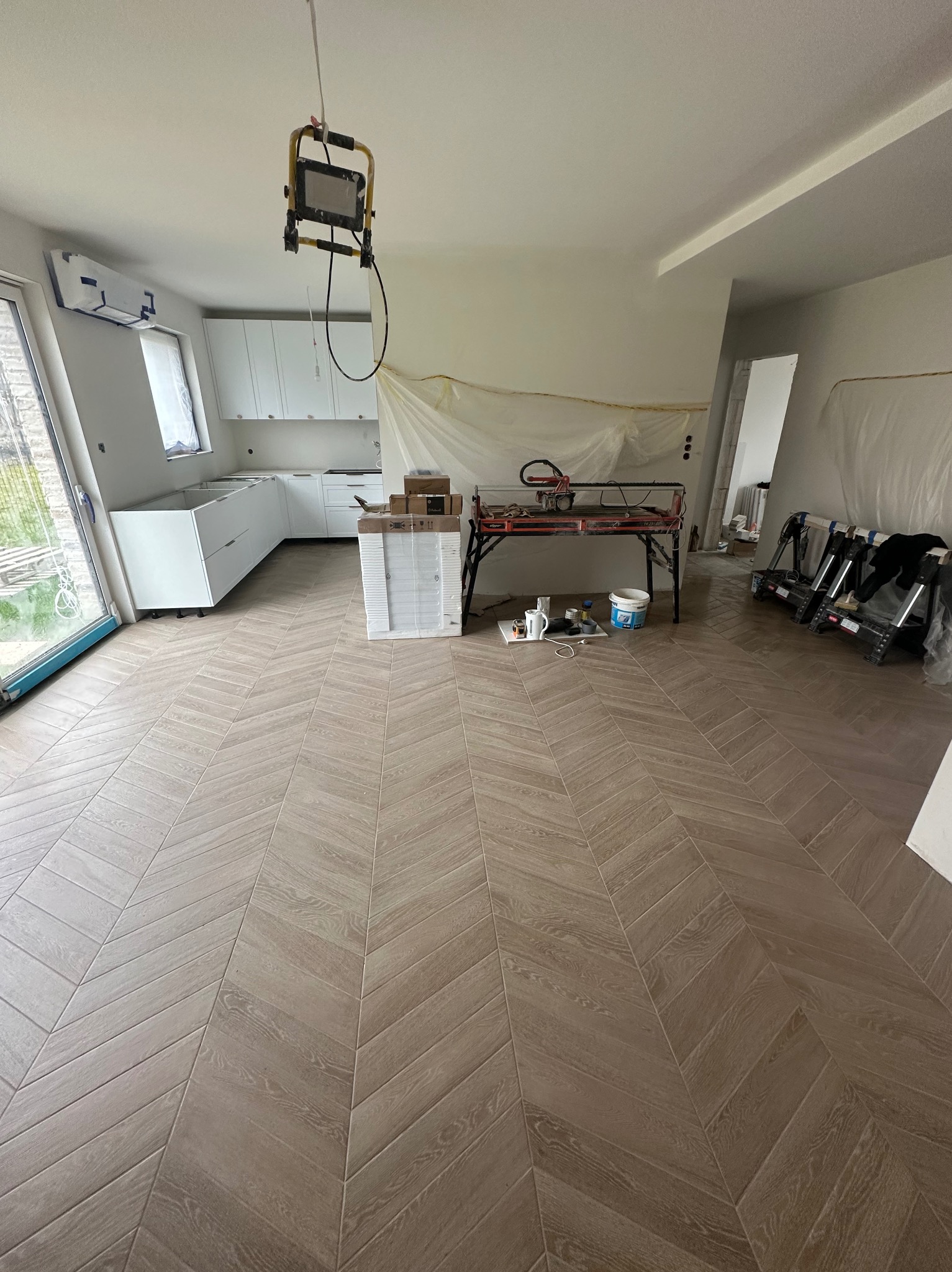 Ułożona podłoga z paneli w jodełkę w jasnym odcieniu drewna, w tle białe, częściowo zamontowane szafki kuchenne i osłonięte folią ściany, widoczny sprzęt do cięcia paneli oraz lampa budowlana.