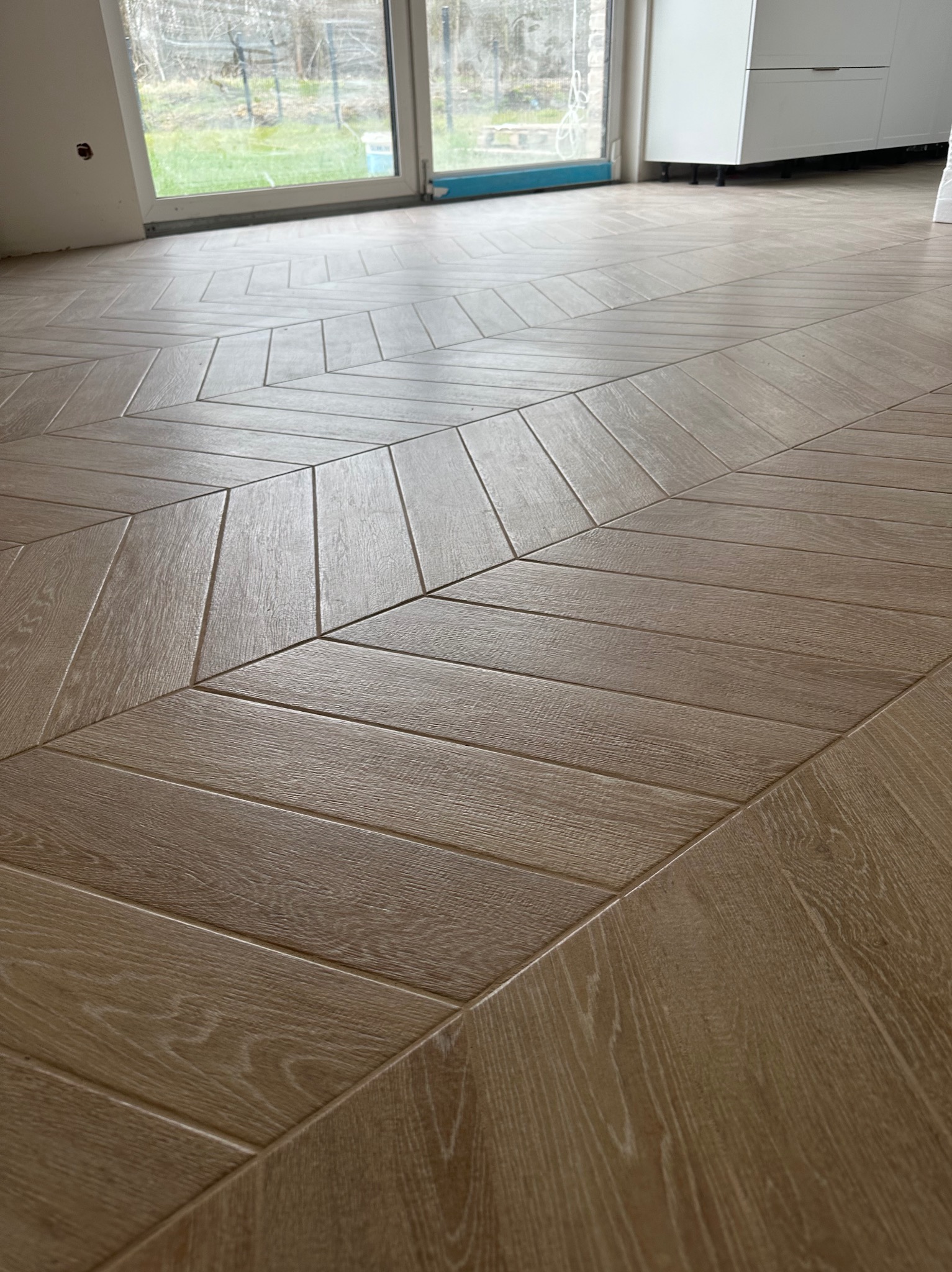 Ułożone panele podłogowe w jodełkę francuską, widoczne łączenia i struktura drewna, w tle okno balkonowe i fragment białej szafki.