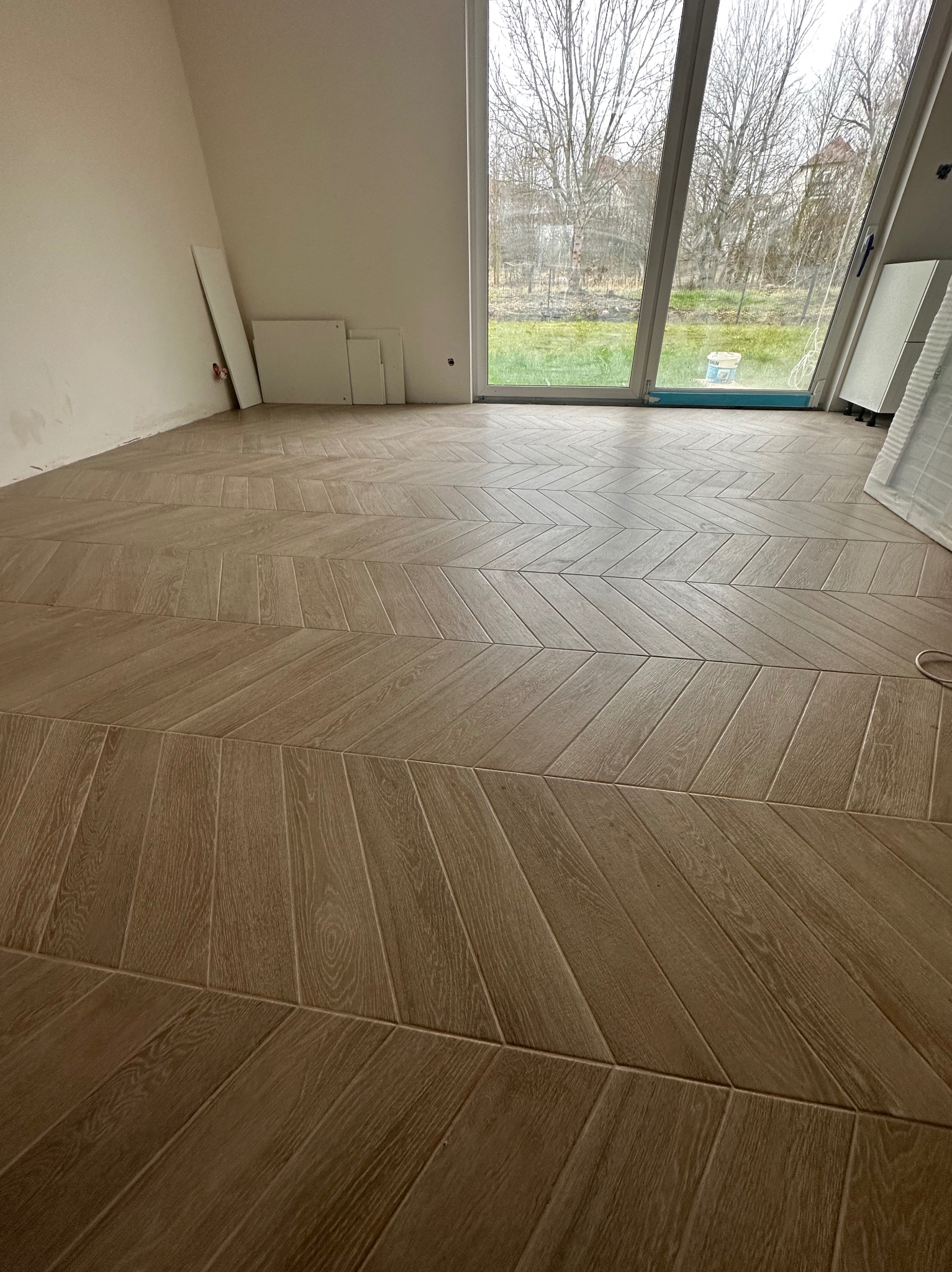 Ułożona pod kątem brązowa podłoga z paneli imitujących drewno w jasnym, surowym wnętrzu z dużym oknem i widokiem na ogród.