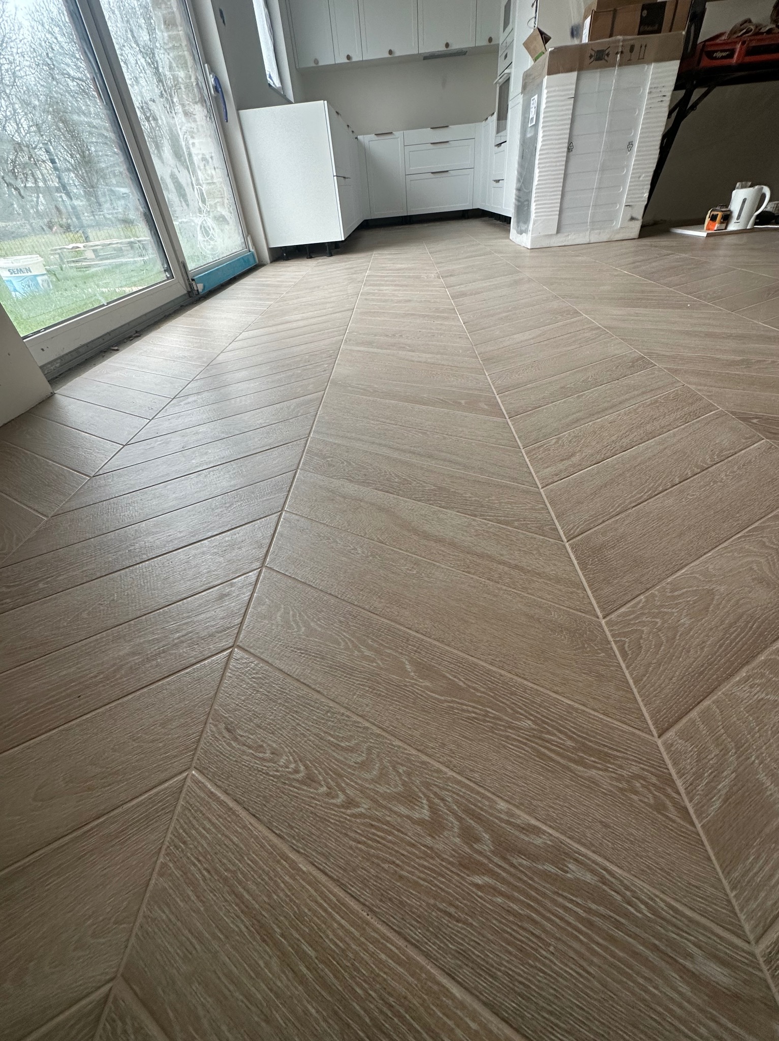 Ułożona pod kątem jasnobrązowa podłoga z paneli imitujących drewno w nowym mieszkaniu, widoczna część białej zabudowy kuchennej i okno z widokiem na zewnątrz.