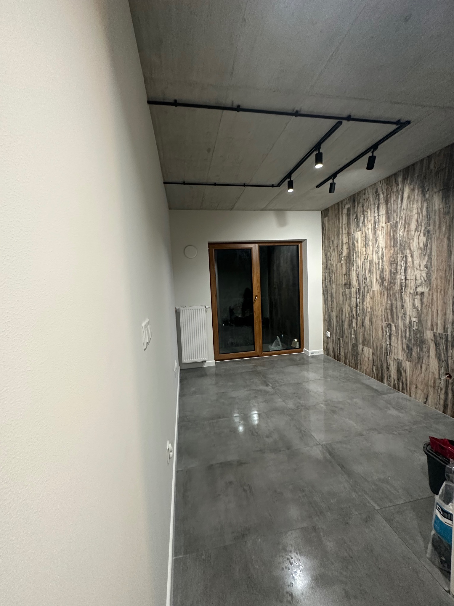 Wnętrze pokoju z wykończoną ścianą z płytek imitujących drewno, szarą podłogą z płytek, białymi ścianami, grzejnikiem, przesuwnymi drzwiami balkonowymi i czarnym oświetleniem szynowym na betonowym...