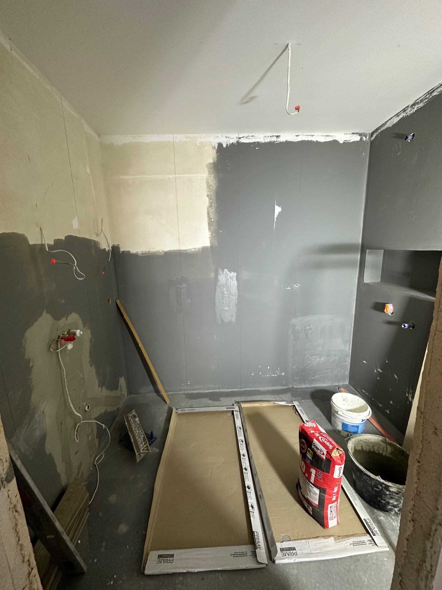 Łazienka w trakcie remontu: częściowo pomalowane ściany na szaro, widoczne przewody elektryczne, paczka kleju Sopro, płytki ceramiczne w opakowaniach i wiadro.