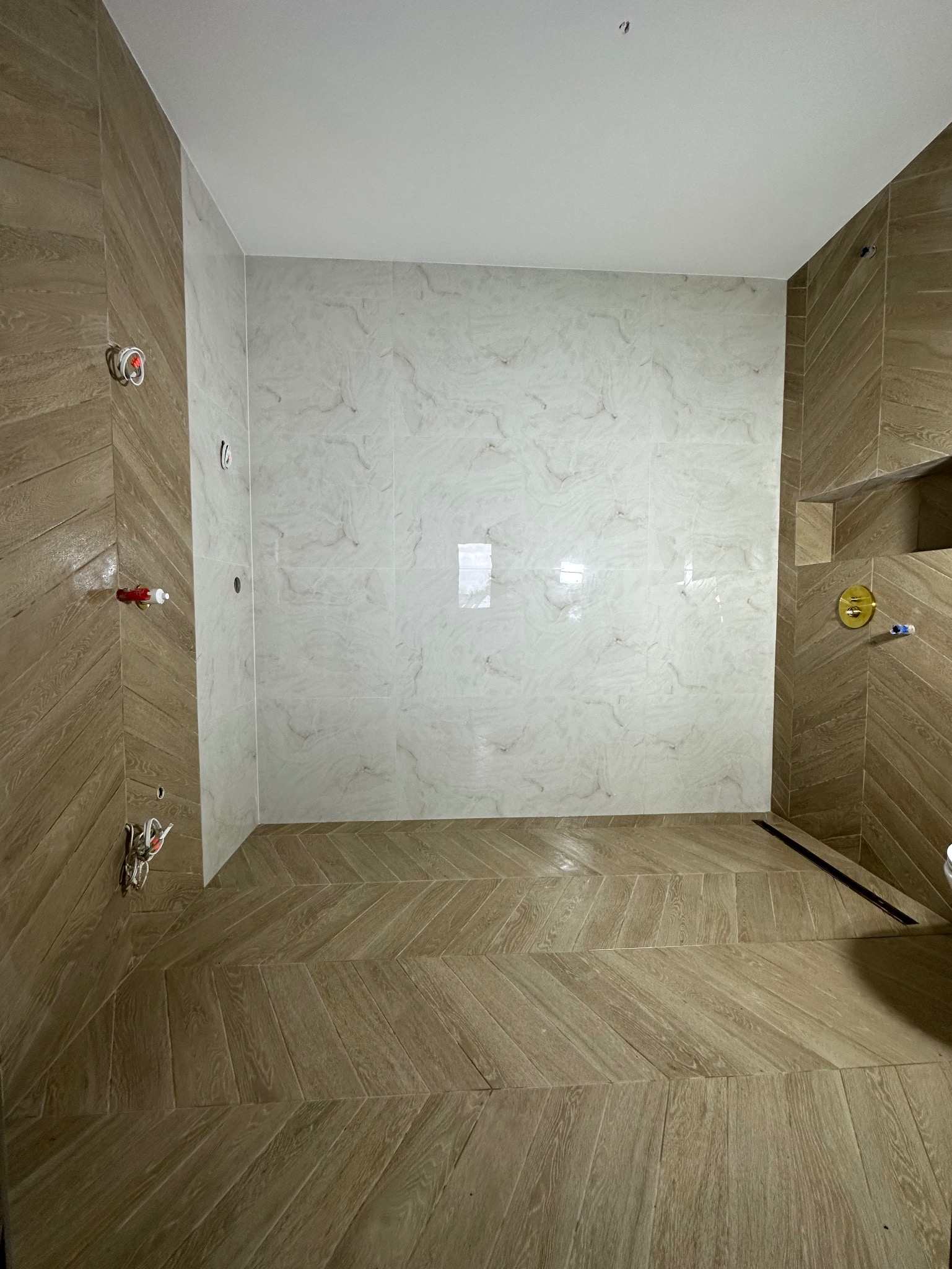 Wnętrze łazienki w trakcie wykańczania, widoczne płytki imitujące drewno ułożone w jodełkę na podłodze i ścianach, białe marmurowe płytki na ścianie naprzeciwko, oraz wystające instalacje...