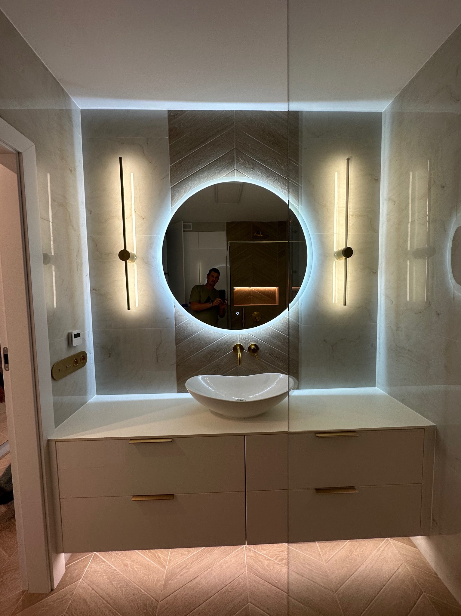 Nowoczesna łazienka z okrągłym lustrem podświetlanym LED, umywalką nablatową, szafką z szufladami i złotymi akcentami, widoczny fragment podłogi z parkietem ułożonym w jodełkę.