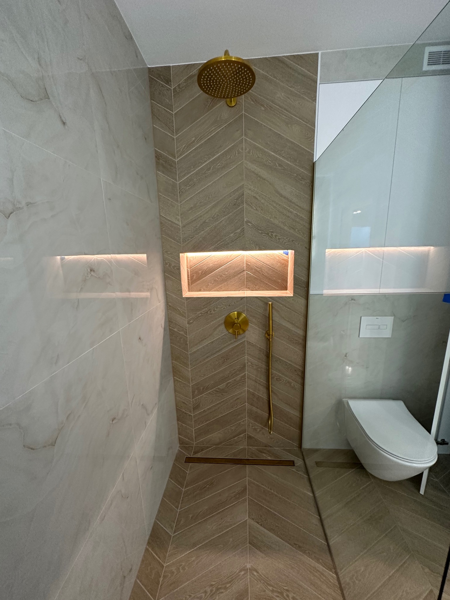 Nowoczesna łazienka z kabiną prysznicową typu walk-in, wykończona płytkami imitującymi drewno ułożonymi w jodełkę na ścianie prysznica i podłodze, złota deszczownica i bateria, podświetlana wnęka...