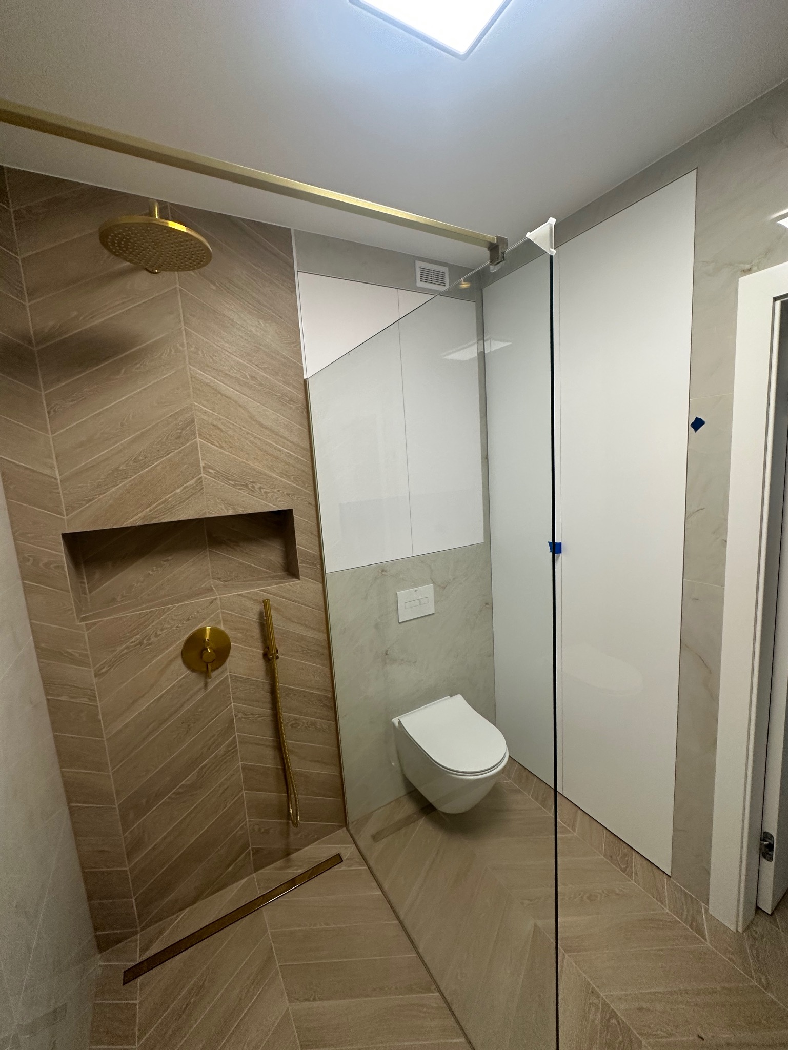 Nowoczesna łazienka z kabiną prysznicową typu walk-in, wykończona płytkami ułożonymi w jodełkę, złotą armaturą, odpływem liniowym i podwieszaną toaletą.