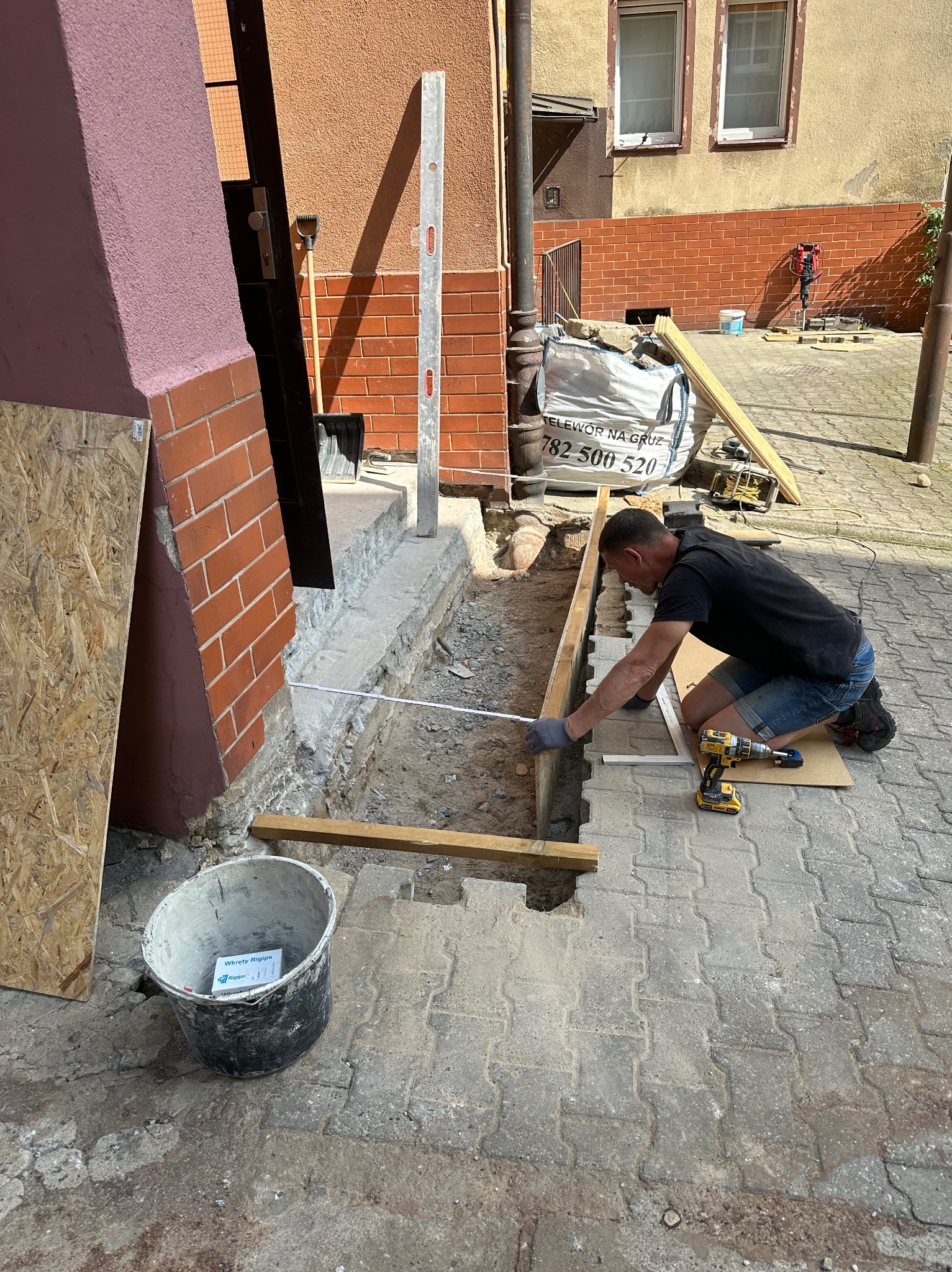 Pracownik w Szczecinie mierzy odległość przed wylaniem betonu pod schody wejściowe do budynku, widoczne narzędzia i materiały budowlane.