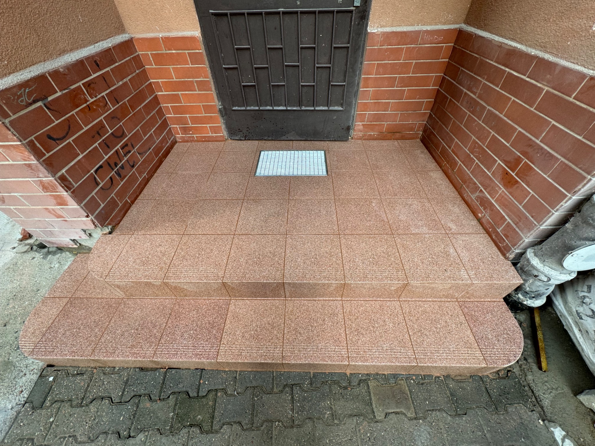 Wyłożone płytkami schody wejściowe do budynku, z widocznymi śladami po graffiti na ceglanej ścianie po lewej stronie.