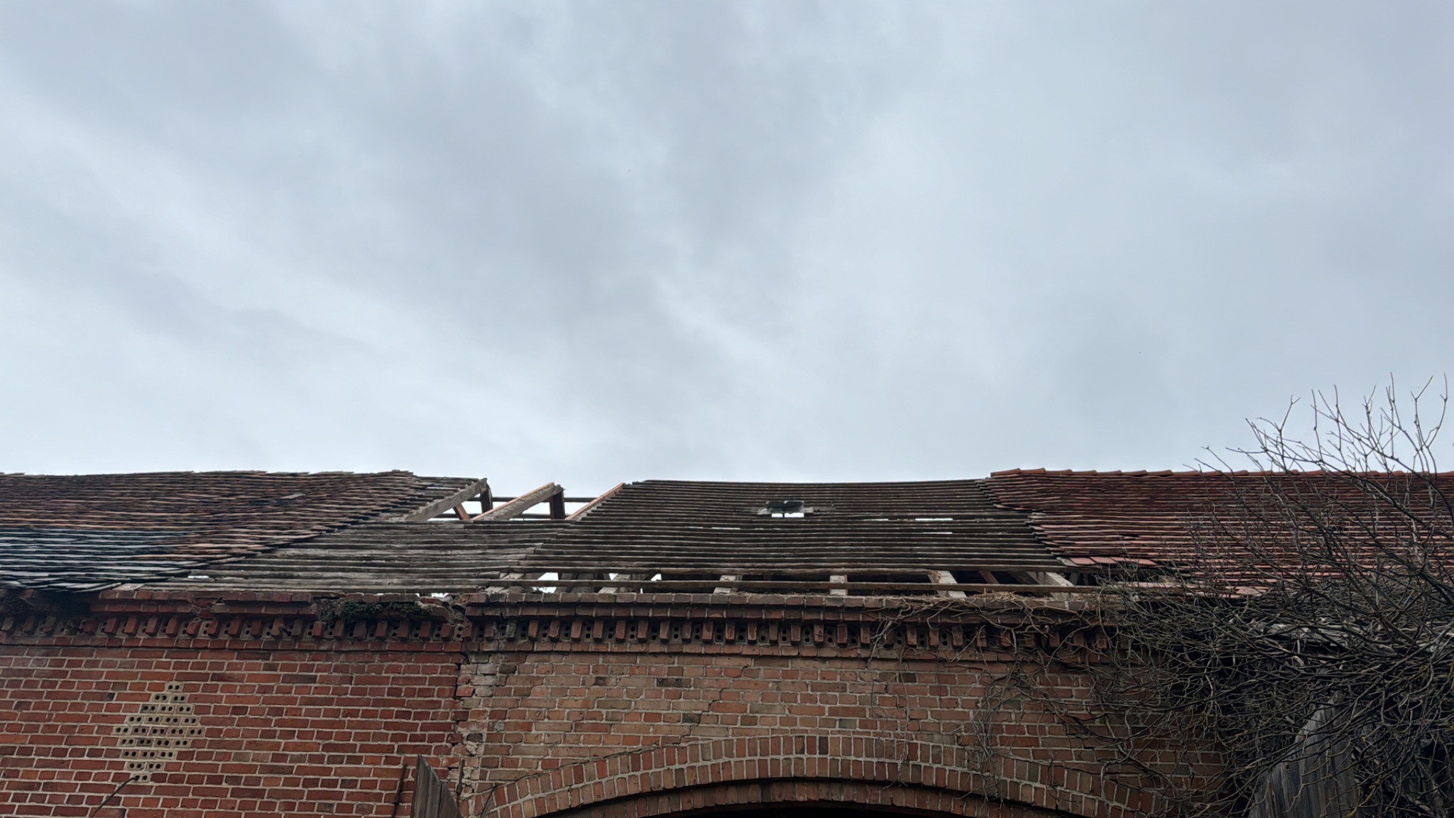Uszkodzony dach z czerwonej dachówki na ceglanym budynku, widoczne fragmenty konstrukcji dachu i dziury w pokryciu, ponure niebo w tle.