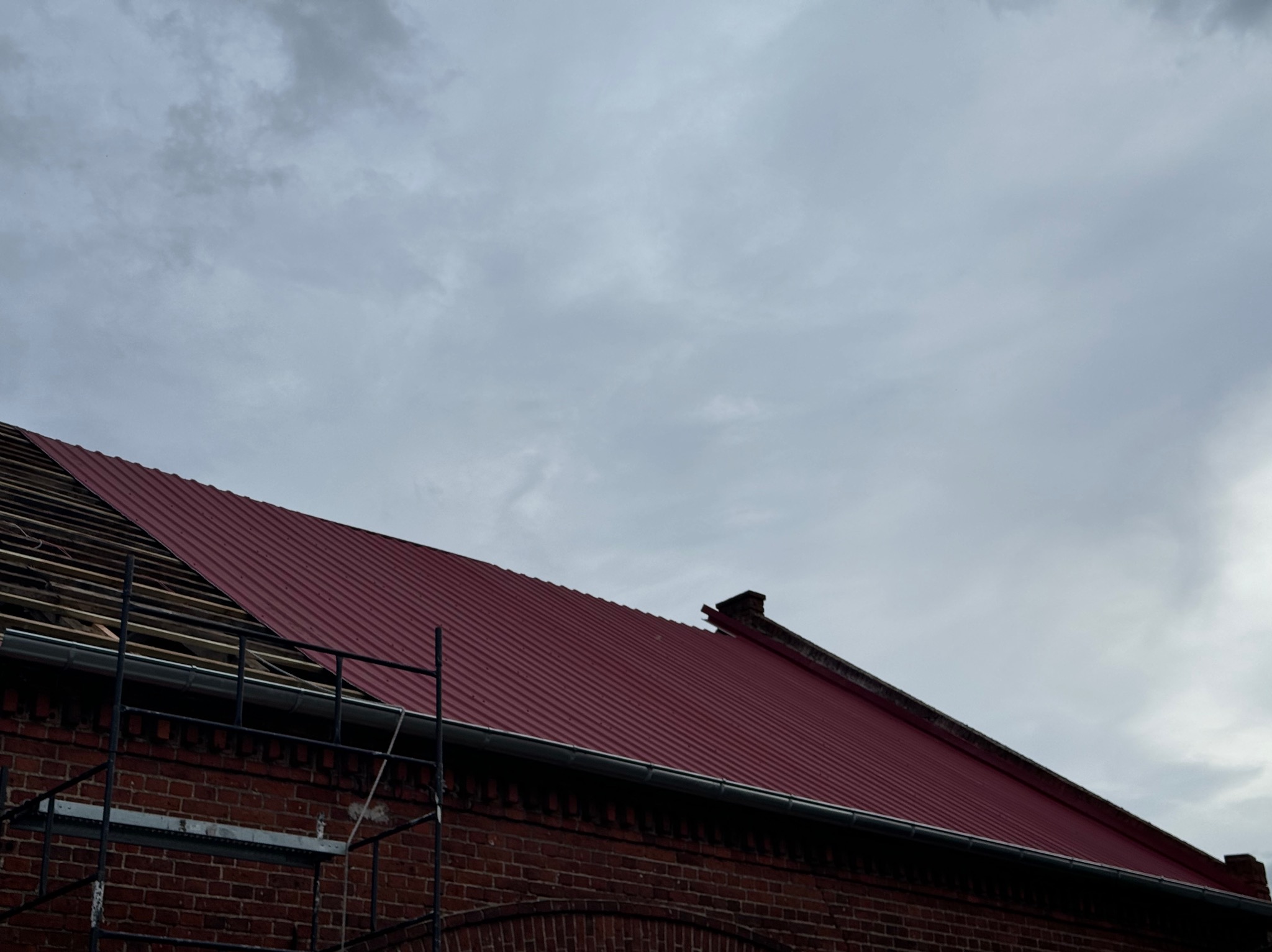 Częściowo wymienione, czerwone, blaszane pokrycie dachu na ceglanym budynku, widoczne rusztowanie i fragment starej konstrukcji dachowej.
