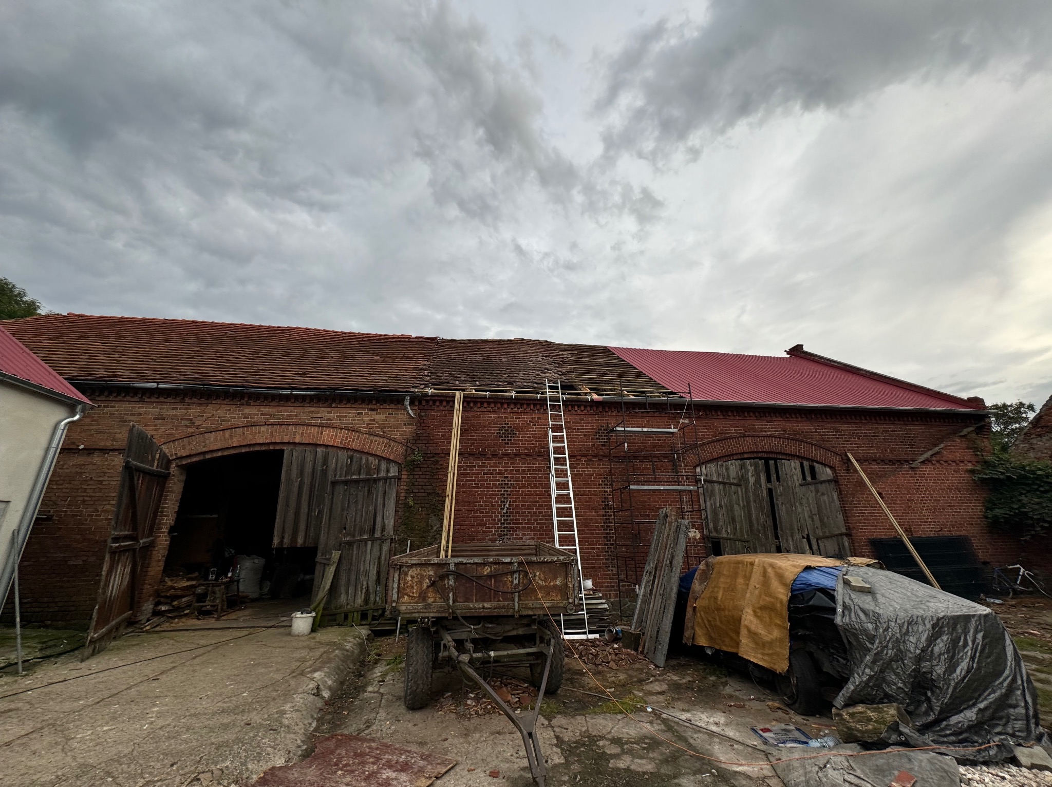 Ceglany budynek gospodarczy z częściowo wymienionym dachem z czerwonej dachówki, drabina oparta o ścianę, przyczepa i materiały budowlane na podwórzu, pochmurne niebo w tle.