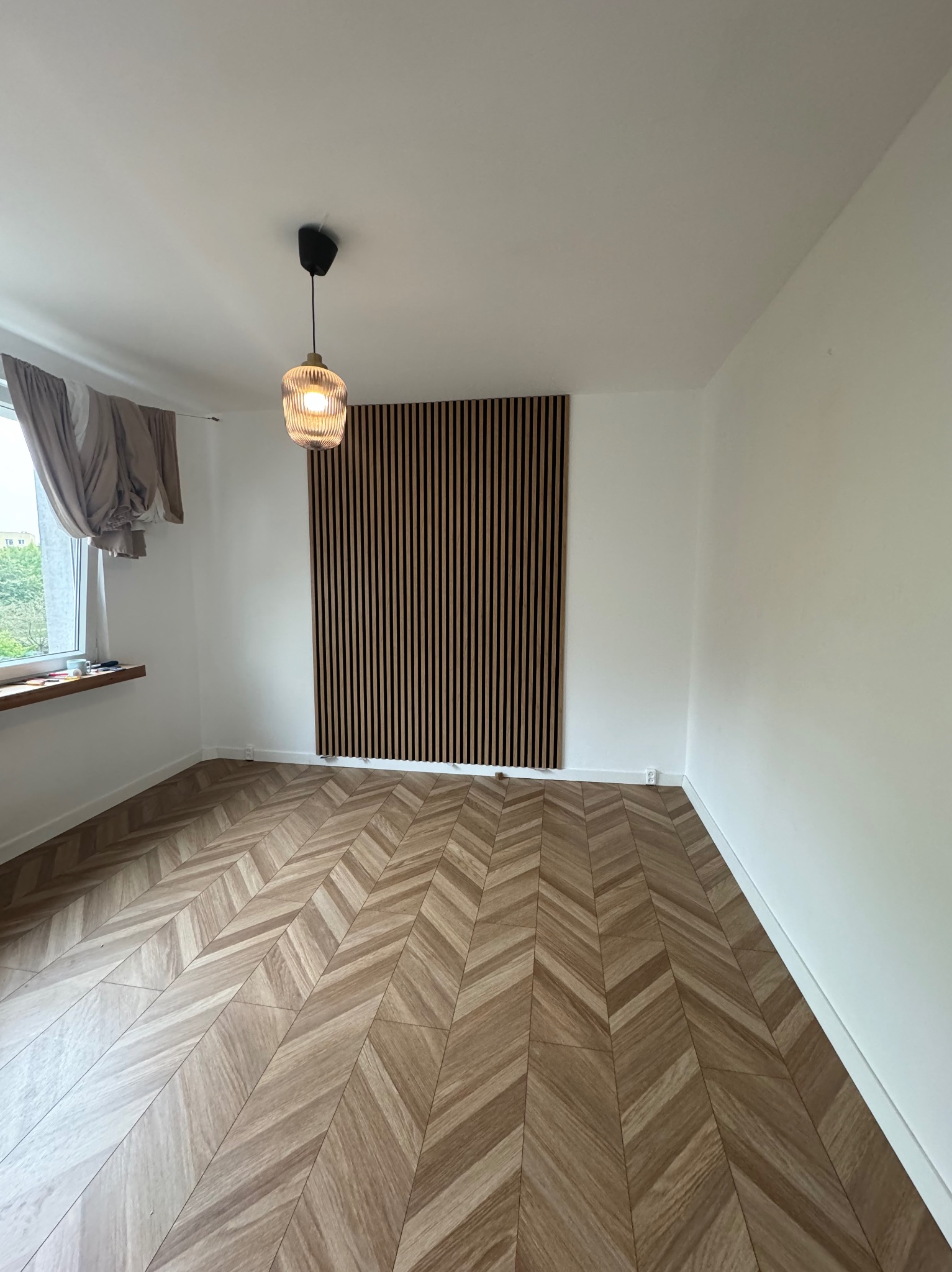 Pokój po remoncie z drewnianą podłogą w jodełkę, drewnianymi lamelami na ścianie i wiszącą lampą z żarówką Edison