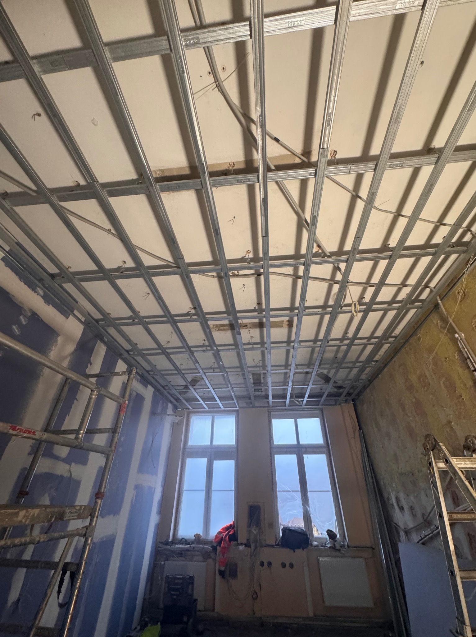 Montaż metalowej konstrukcji sufitu podwieszanego w remontowanym pokoju. Widoczne rusztowania i okna. Prace wykończeniowe w trakcie realizacji.