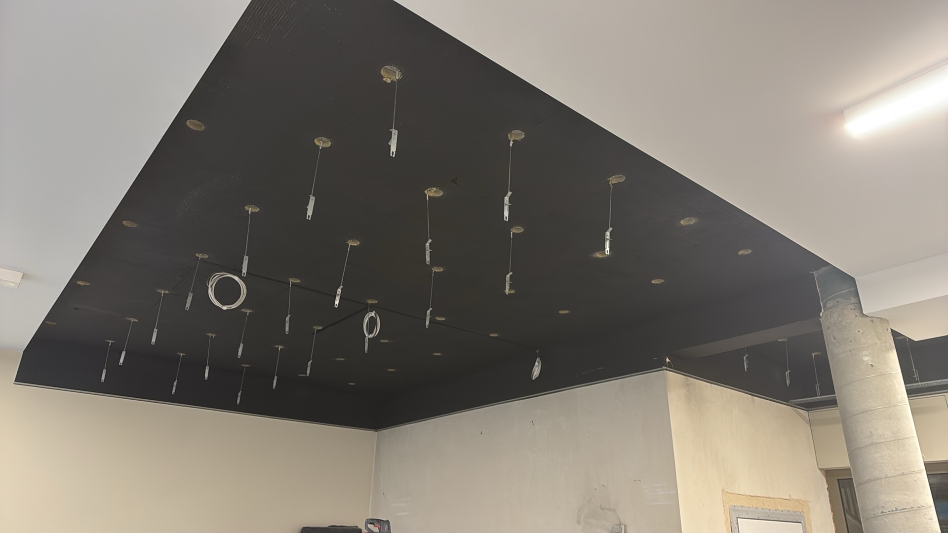 Czarny sufit podwieszany z punktami świetlnymi w trakcie montażu, widoczne przewody i elementy mocujące. Ściany w surowym stanie, kolumna konstrukcyjna.