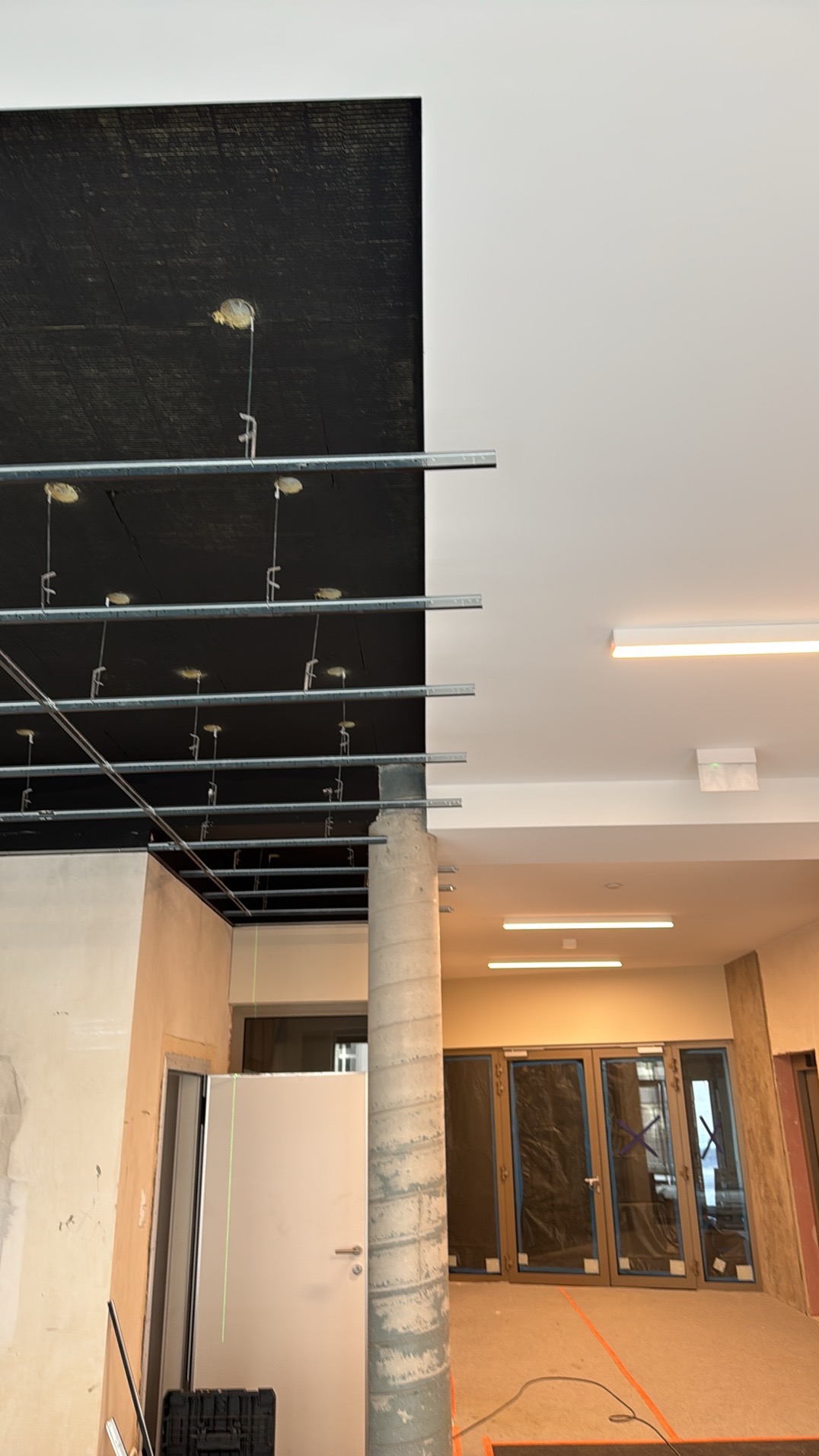 Montaż sufitu podwieszanego z widoczną konstrukcją metalową i instalacją oświetleniową w nowym budynku. Widoczne drzwi i okna.