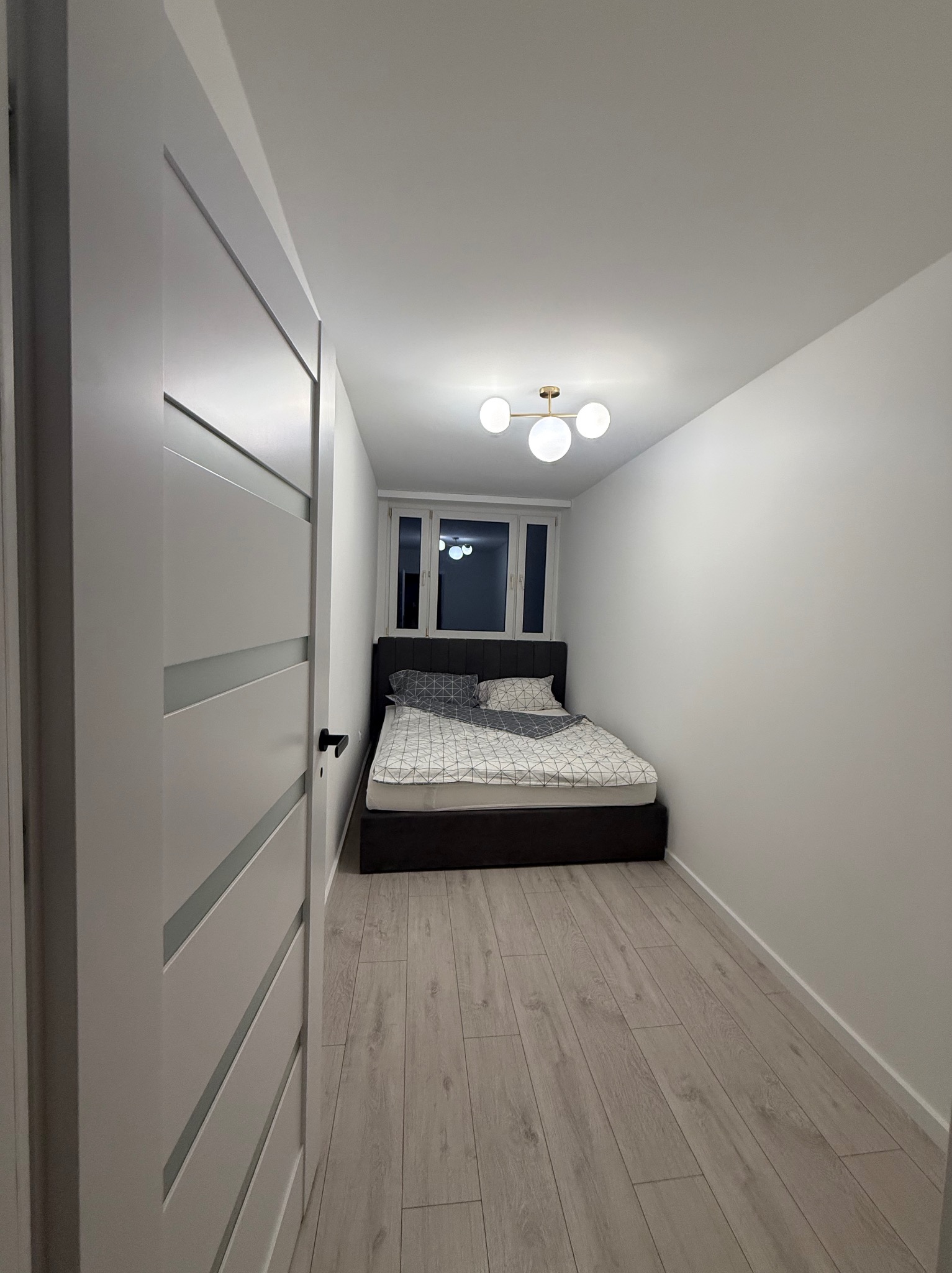 Wnętrze małej sypialni po remoncie: białe ściany, drewniana podłoga, nowoczesne drzwi z przeszkleniami i łóżko pod oknem. Minimalistyczny design.