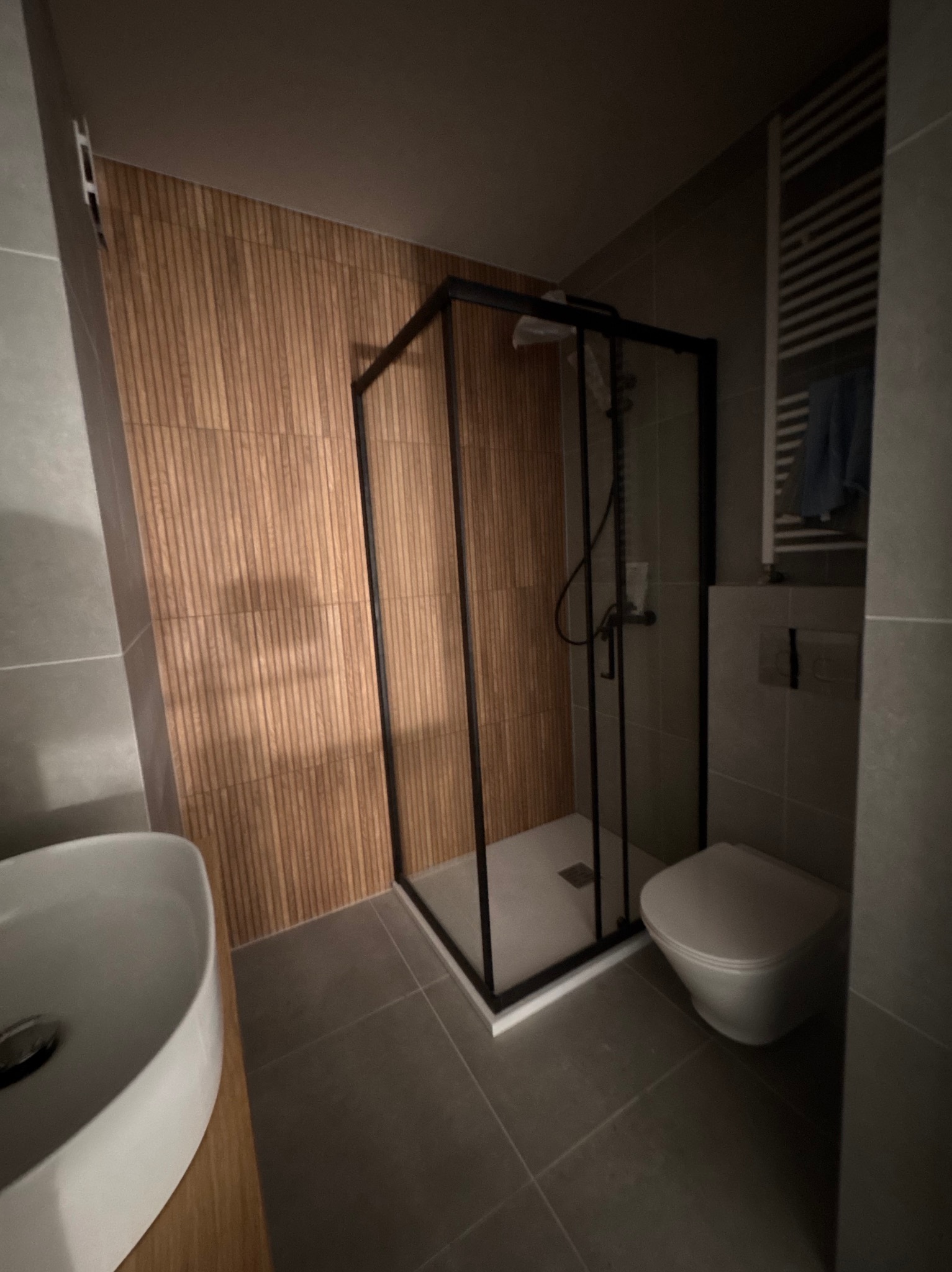 Nowoczesna łazienka z kabiną prysznicową w czarnej ramie, umywalką nablatową i drewnopodobną okładziną ściany. Minimalistyczny design, szare płytki podłogowe.