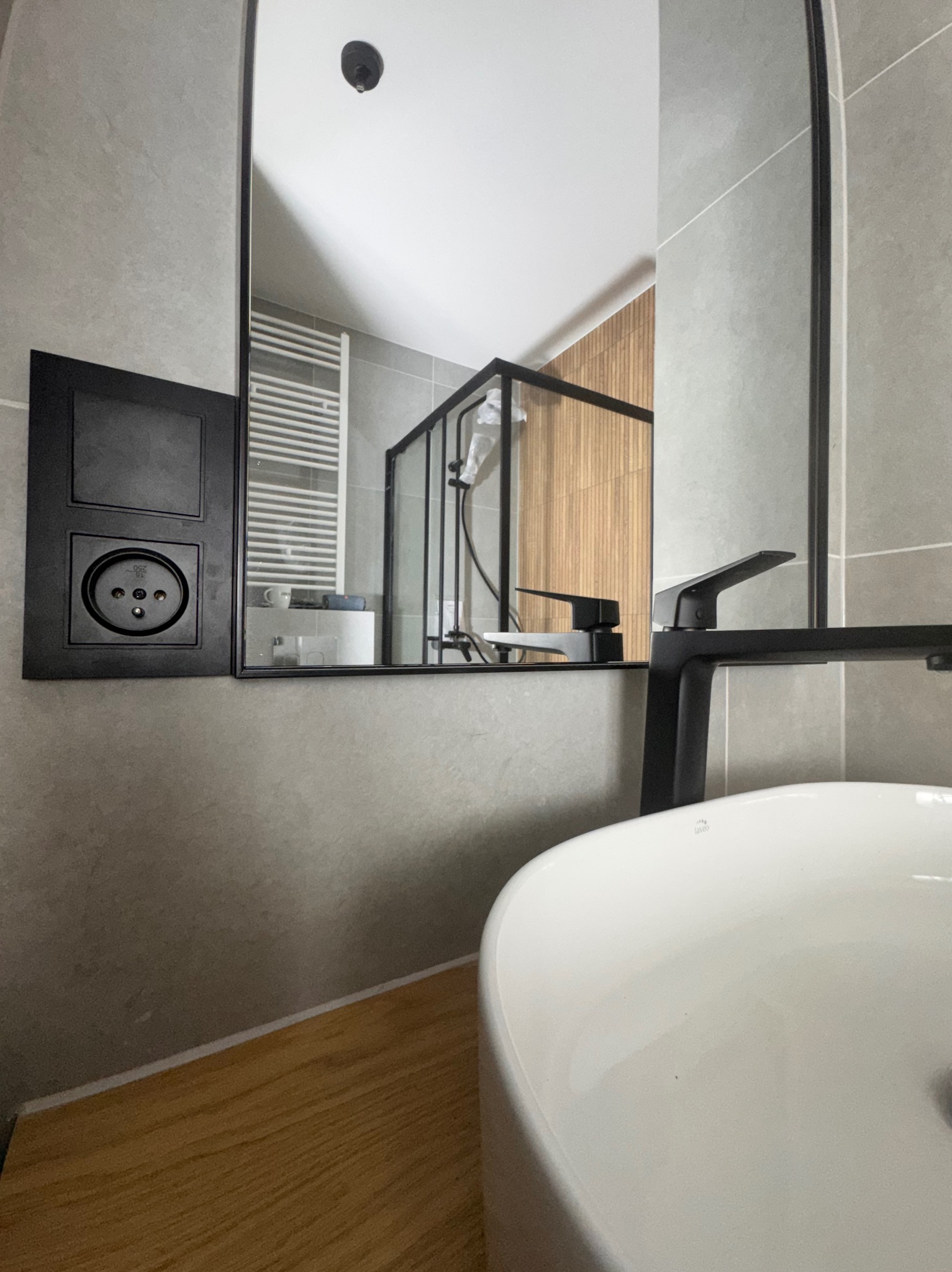 Nowoczesna łazienka z szarą płytką, czarną armaturą i drewnianym blatem. W lustrze odbija się kabina prysznicowa z drewnianymi panelami. Widoczny kontakt elektryczny.