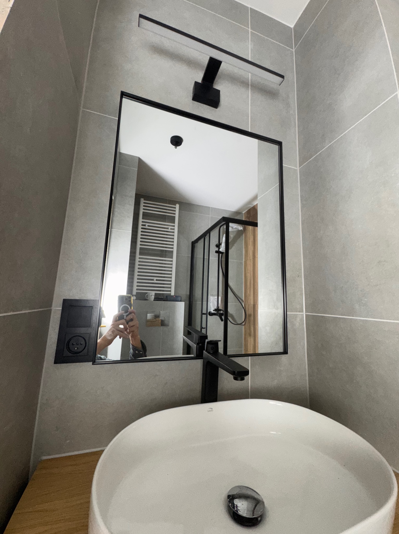 Nowoczesna łazienka z umywalką nablatową, czarną armaturą i lustrem w czarnej ramie. W tle widoczny grzejnik drabinkowy i kabina prysznicowa.