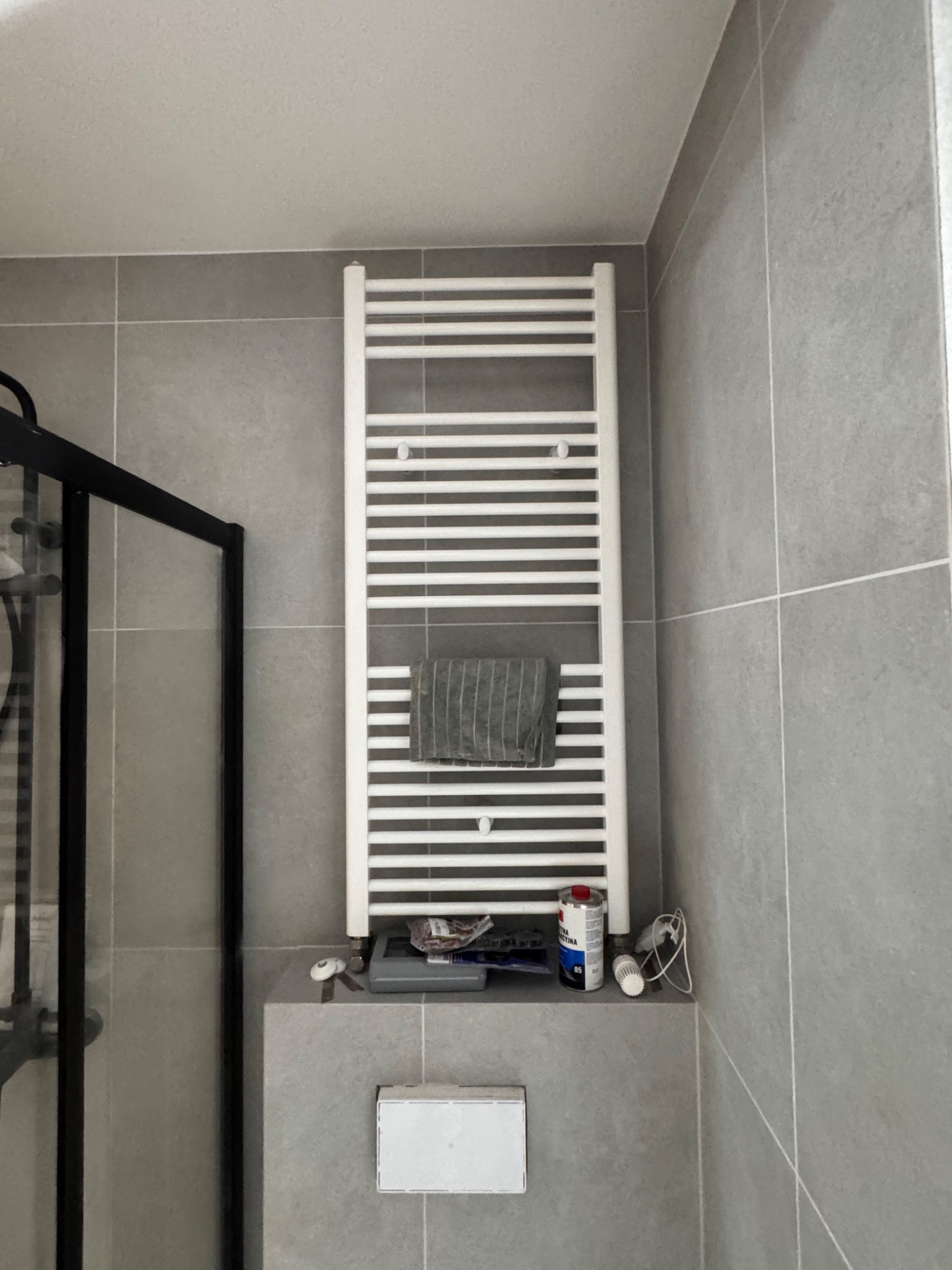 Szara łazienka z grzejnikiem drabinkowym, na którym wisi ręcznik. Na półce pod grzejnikiem leżą narzędzia i puszka farby. Fragment kabiny prysznicowej po lewej.