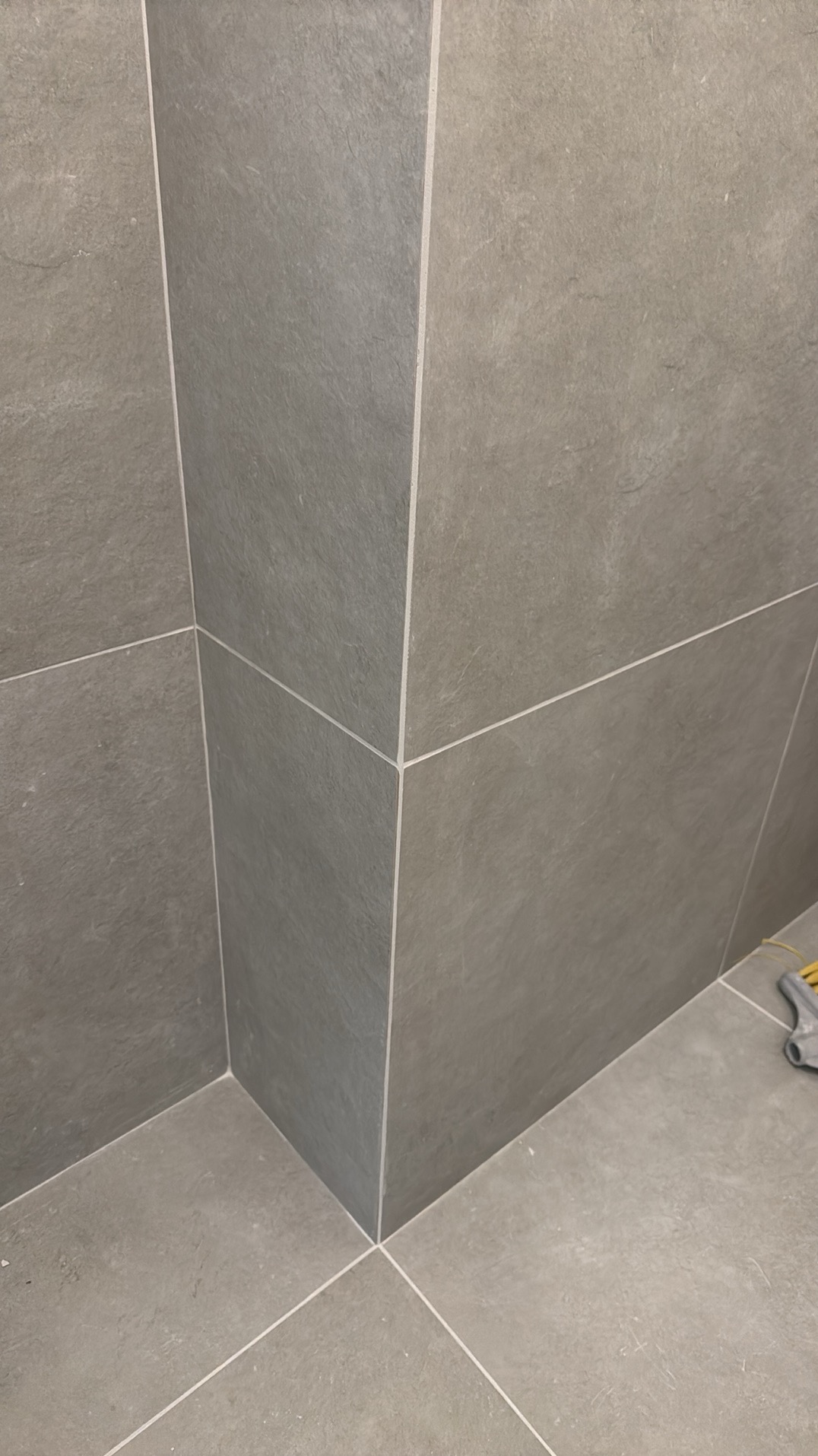 Szare, matowe płytki w rogu pomieszczenia, ułożone z precyzją i cienką fugą. Fragment narzędzia w prawym dolnym rogu sugeruje prace wykończeniowe.
