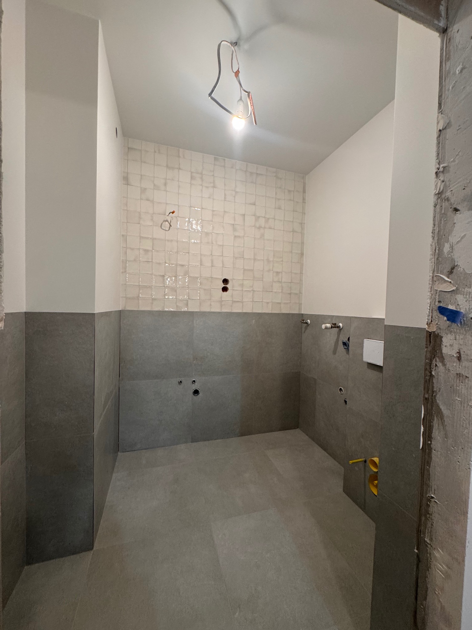 Wnętrze łazienki w trakcie prac wykończeniowych: ułożone płytki ścienne do połowy wysokości, widoczne instalacje wodne, surowy beton przy framudze drzwi.