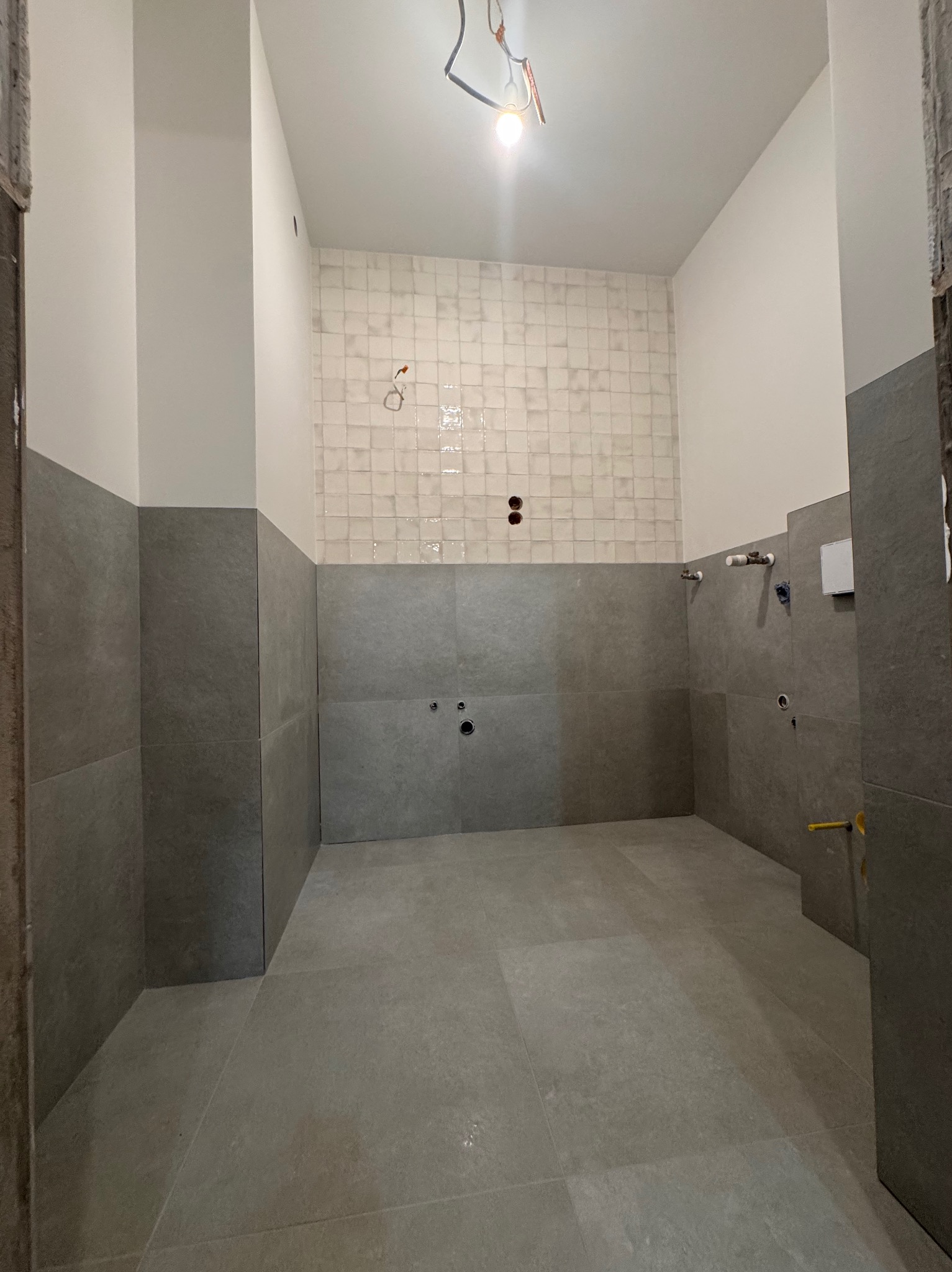 Wykończona łazienka z szarymi płytkami do połowy ściany i białymi kwadratowymi płytkami powyżej. Widoczne instalacje wodne i elektryczne. Minimalistyczny design.