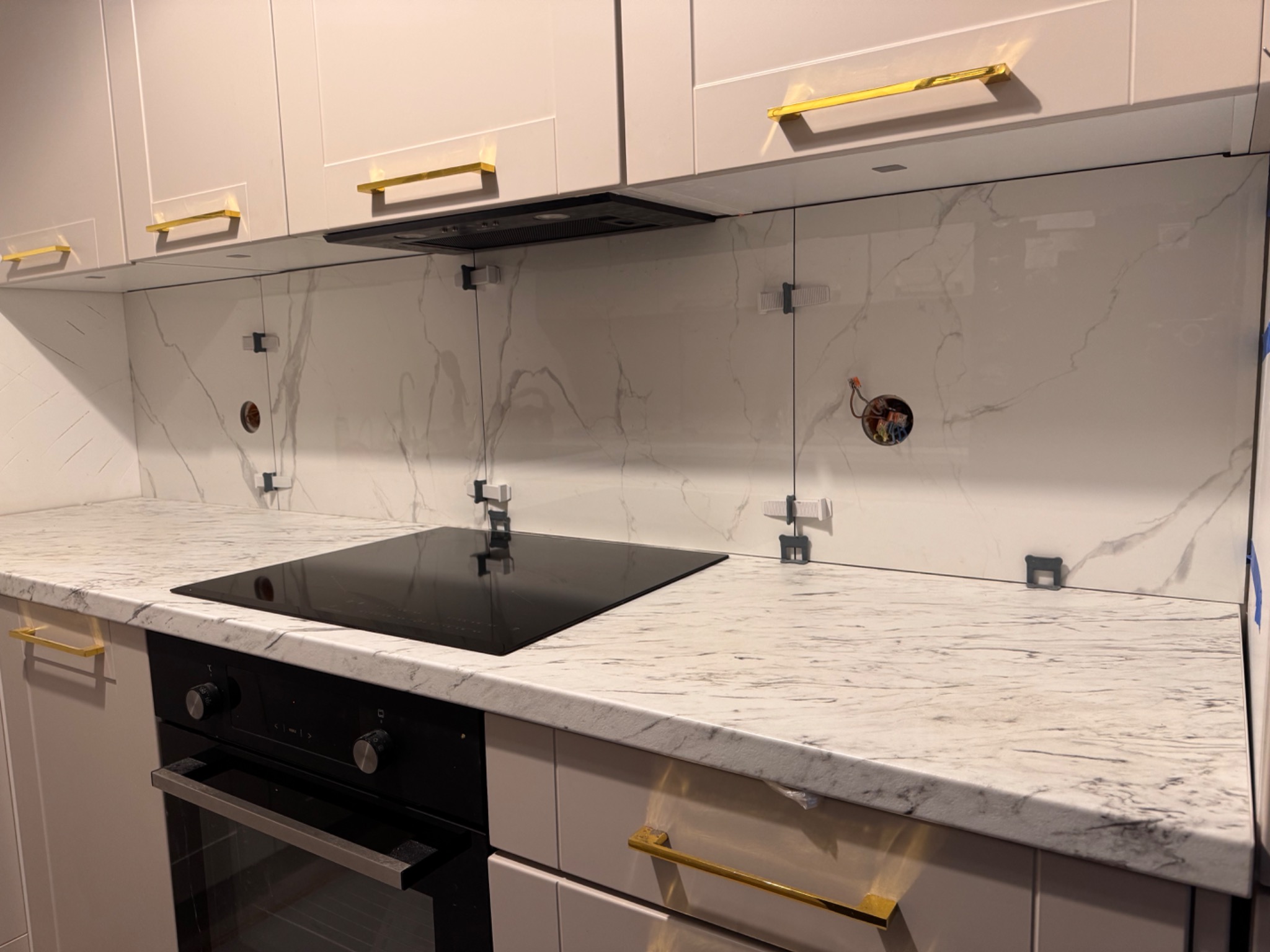 Montaż paneli ściennych imitujących marmur w kuchni, widoczne klipsy montażowe, płyta indukcyjna i piekarnik w zabudowie. Złote uchwyty szafek dodają elegancji.