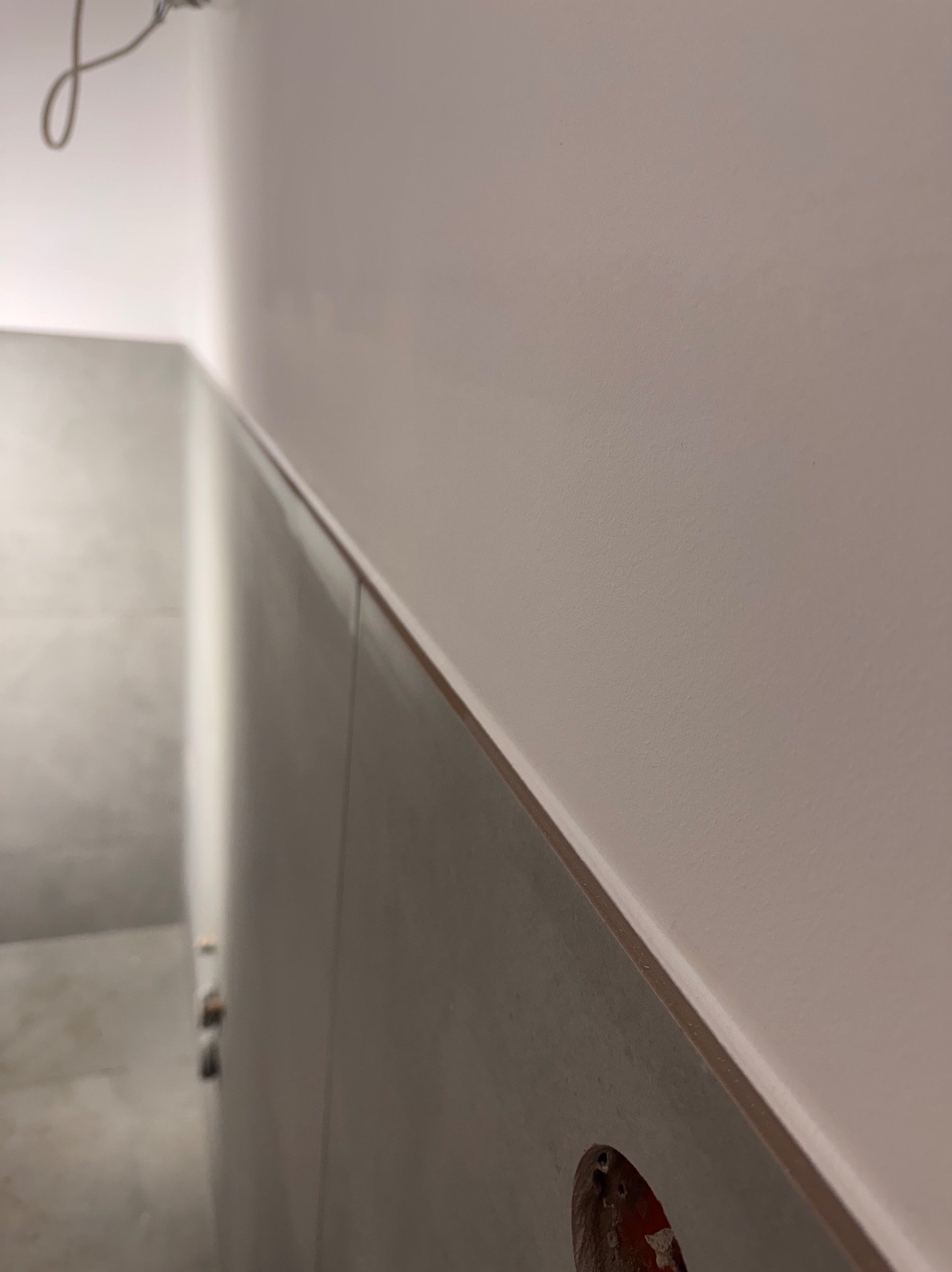 Ściana łazienki z szarymi płytkami do połowy wysokości i pomalowaną górną częścią na jasny kolor. Widoczne gniazdko elektryczne.