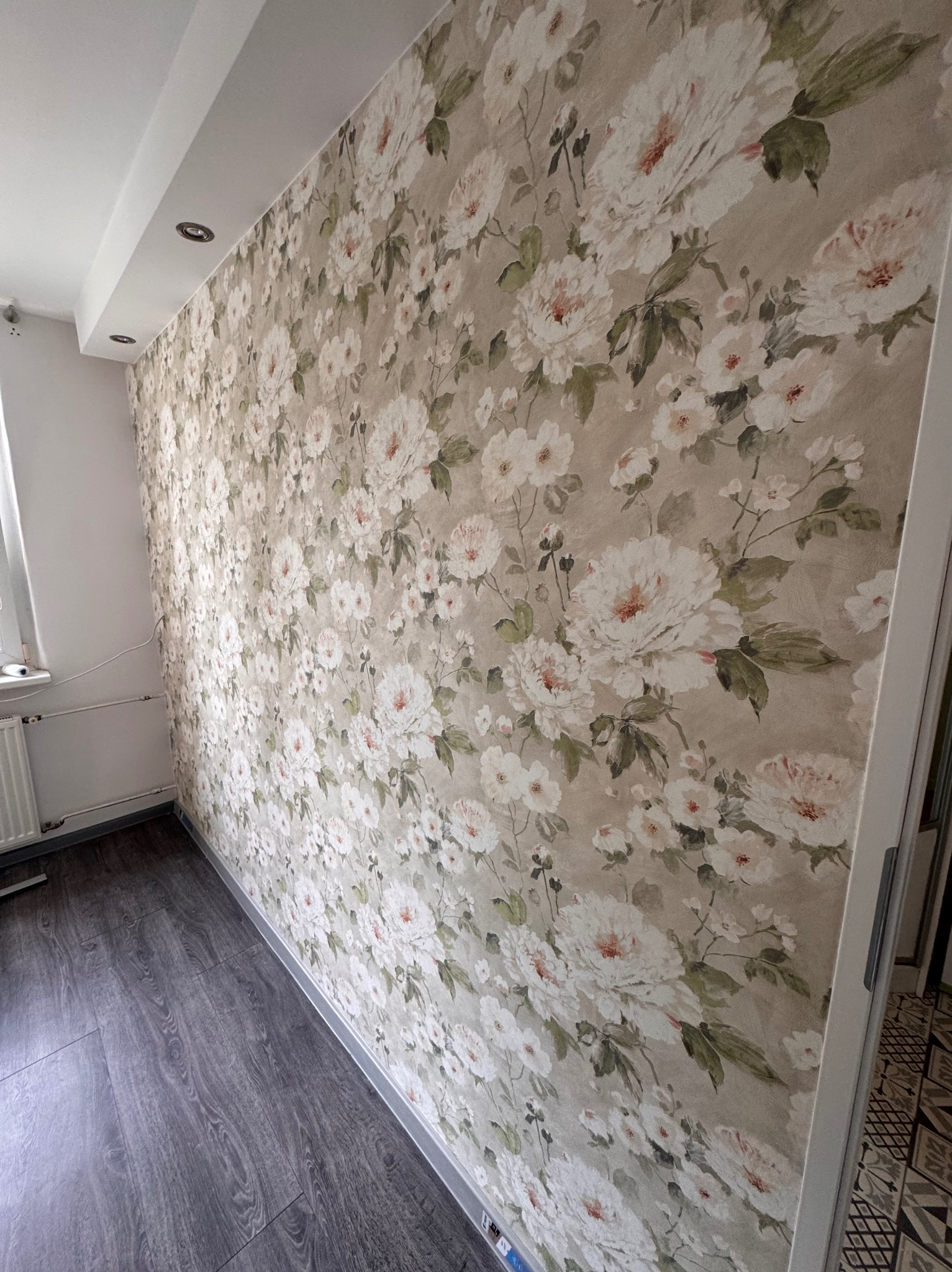 Pokój po tapetowaniu ścian w kwiaty i ułożeniu ciemnej podłogi. Widoczne listwy przypodłogowe i fragment okna. Dekoracyjne wykończenie wnętrza.