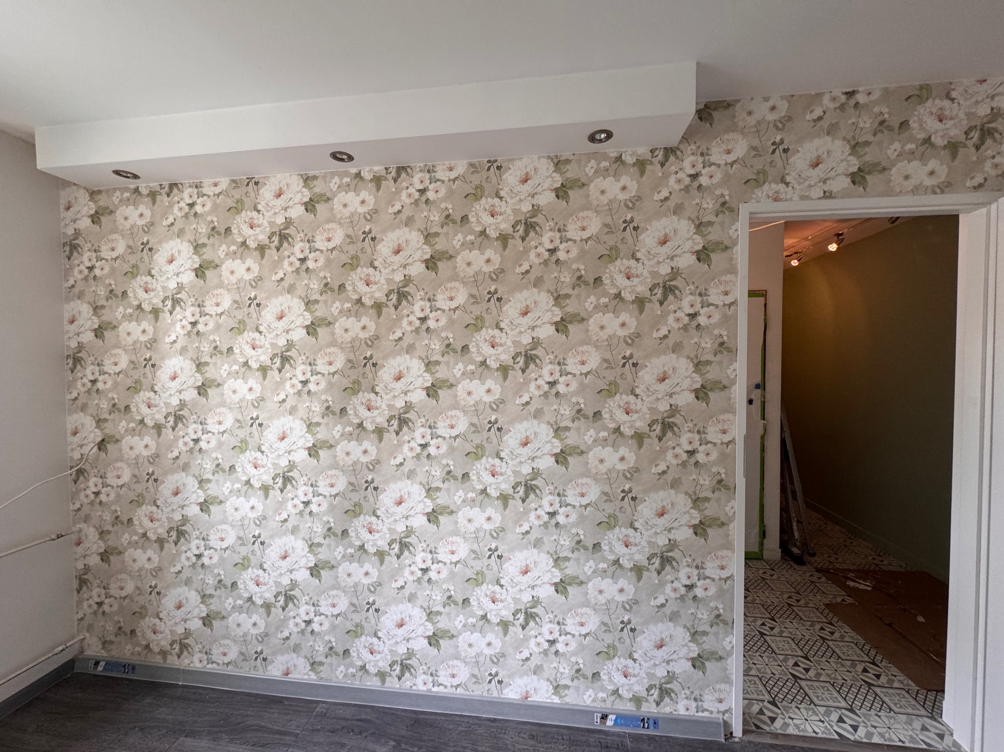 Ściana z tapetą w kwiaty w jasnym pokoju. Widoczny fragment sufitu z oświetleniem punktowym i fragment podłogi. W tle widoczne wejście do innego pomieszczenia.