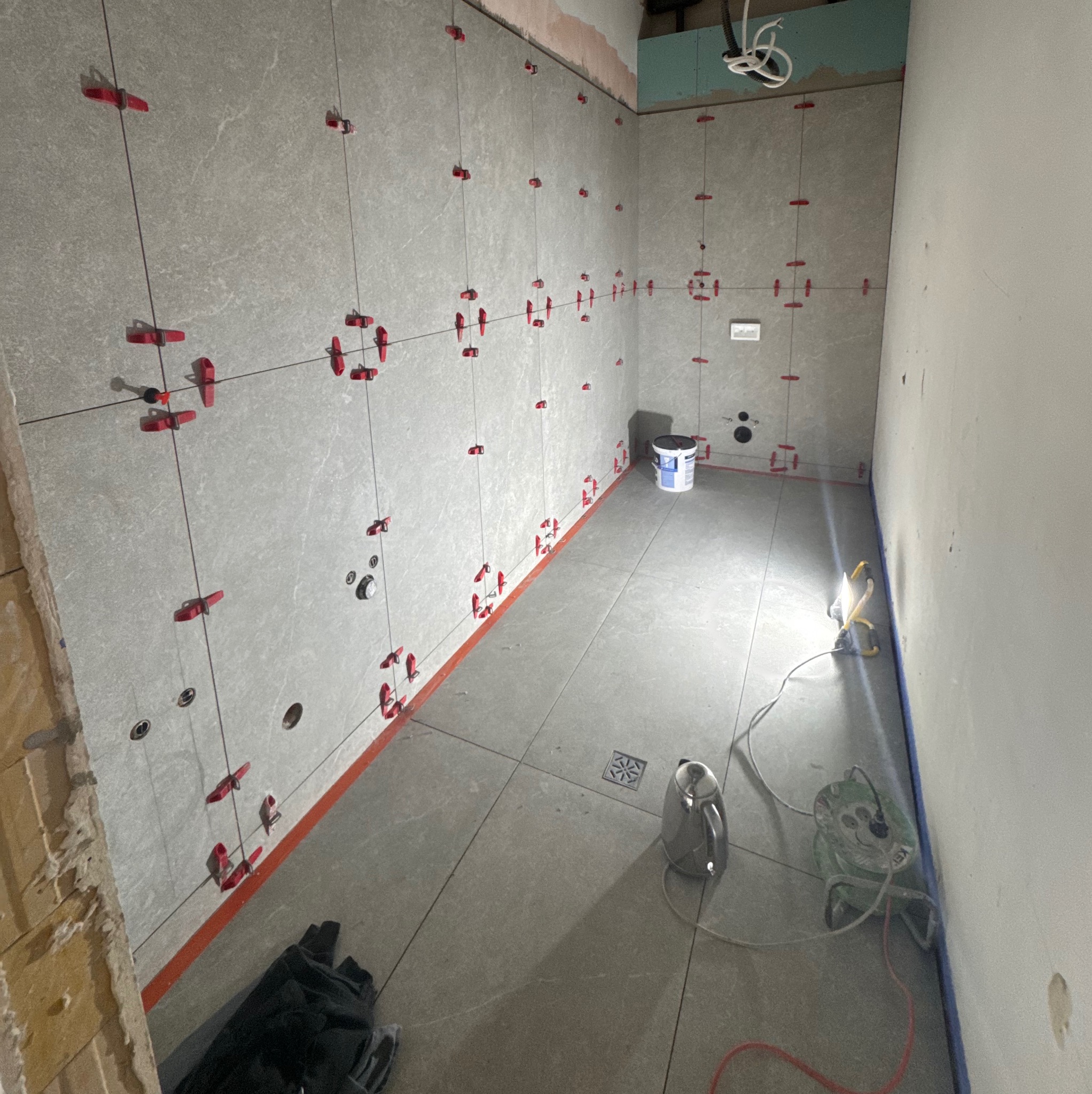 Szare płytki na ścianach i podłodze, system poziomowania płytek z czerwonymi klipsami, widoczne narzędzia glazurnicze, etap prac w łazience.