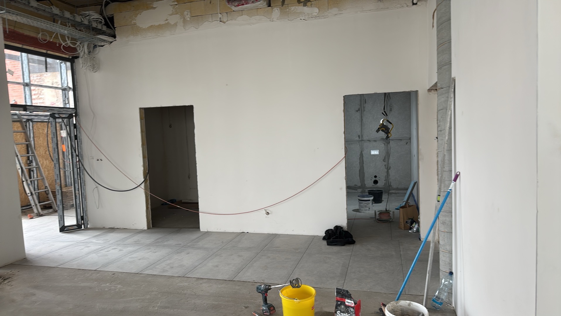Wnętrze w trakcie prac wykończeniowych: położona szara podłoga, ściany przygotowane do malowania, widoczne narzędzia i materiały budowlane. Dwa otwory drzwiowe.