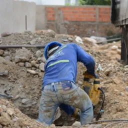 Pracownik w niebieskiej bluzie i kasku używa młota udarowego w wykopie ziemnym, widoczny kabel zasilający i fragment skrzynki elektrycznej.