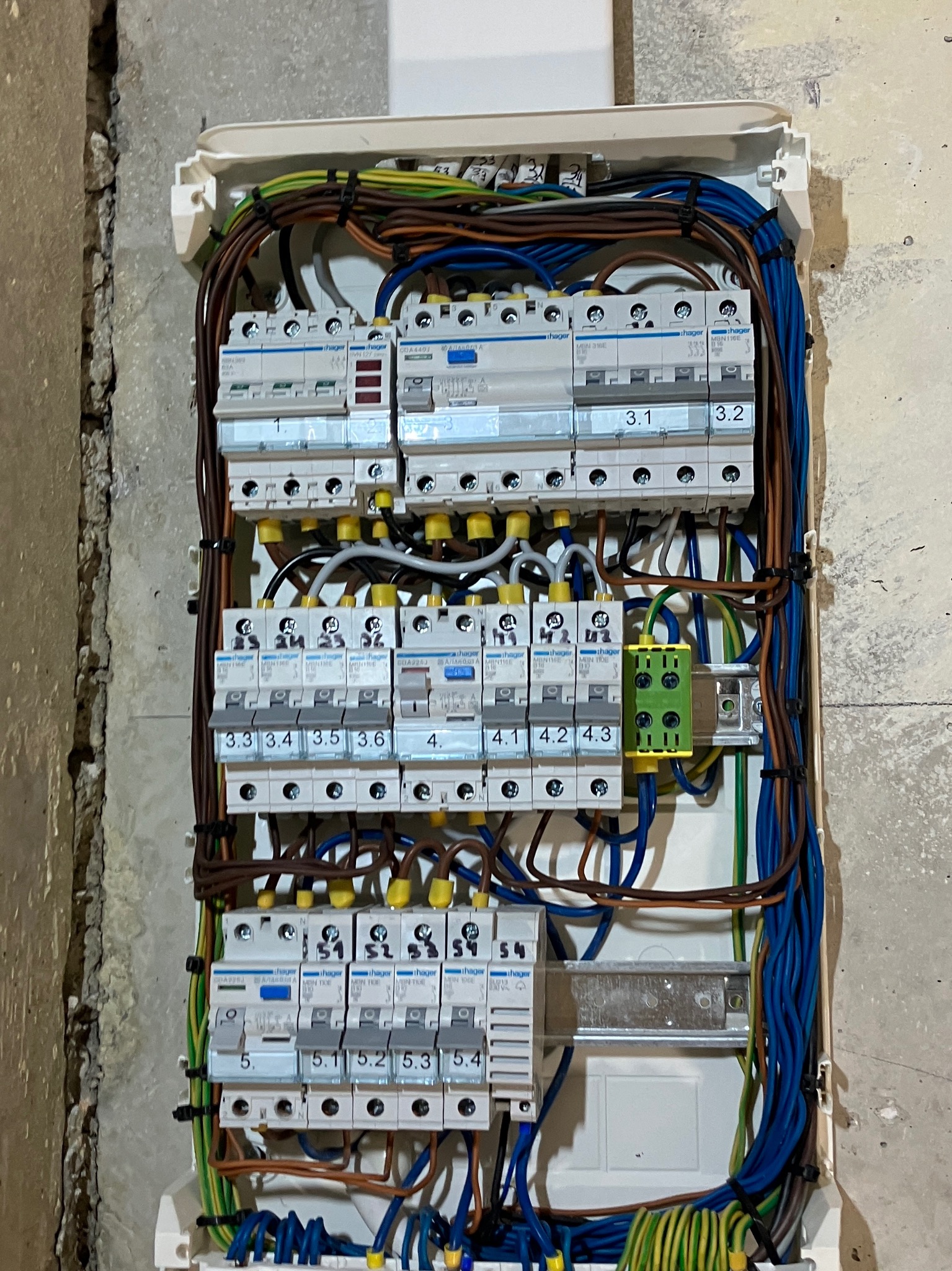 Rozdzielnica elektryczna Hager z zainstalowanymi bezpiecznikami i okablowaniem, widoczna na tle surowej ściany z betonu.