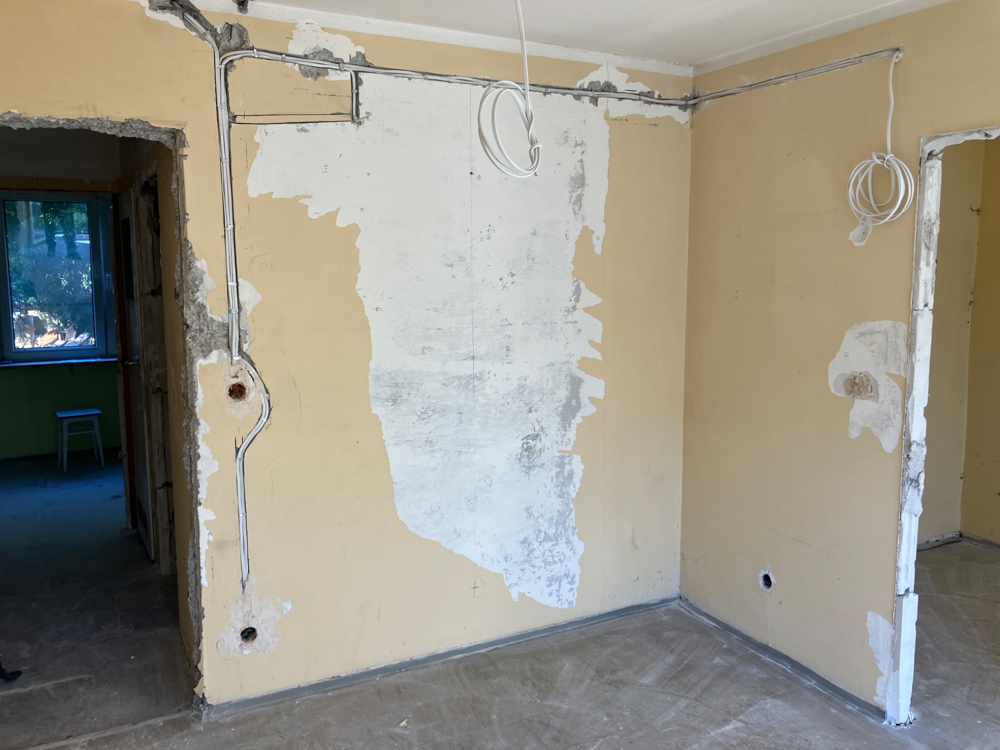 Wnętrze w trakcie remontu: ściany w odcieniach beżu z fragmentami odsłoniętego tynku, widoczne przewody elektryczne w peszlach na ścianie, puszki elektryczne, podłoga wylewka, otwór drzwiowy...