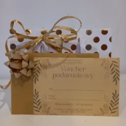 Złoty voucher podarunkowy na masaż od Studio Masażu Anna Konopka, umieszczony w ozdobnej torebce prezentowej w białe i złote kropki, przewiązany złotą wstążką.