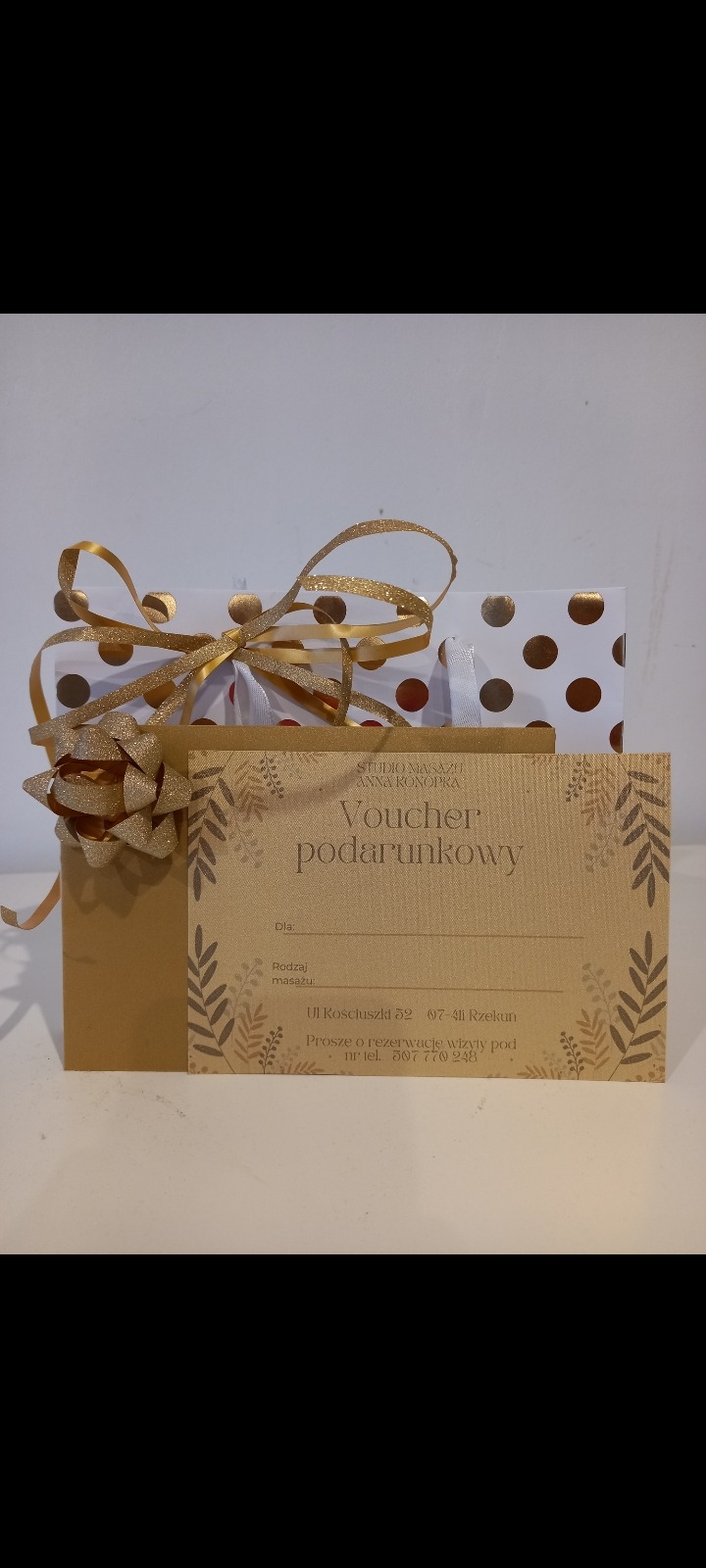 Złoty voucher podarunkowy na masaż od Studio Masażu Anna Konopka, umieszczony w ozdobnej torebce prezentowej w białe i złote kropki, przewiązany złotą wstążką.