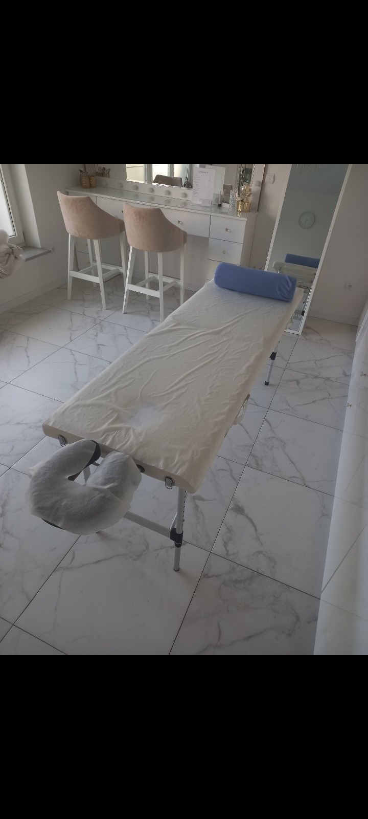 Stół do zabiegów z poduszką pod głowę i wałkiem pod kark, przykryty prześcieradłem, ustawiony w jasnym pomieszczeniu z toaletką i lustrem.