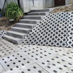 Schody z szarych płyt prowadzące do drzwi wejściowych, obok ażurowa konstrukcja z betonowych elementów i ułożona nawierzchnia z płyt z otworami.