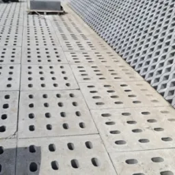 Ułożone betonowe płyty ażurowe na pochyłym terenie, z widocznym w tle fragmentem budynku i wózkiem transportowym.