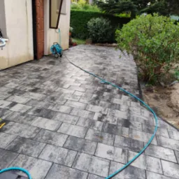 Szara kostka brukowa ułożona przed domem, z widocznym wężem ogrodowym i roślinnością w tle, z otwartymi drzwiami wejściowymi do budynku.